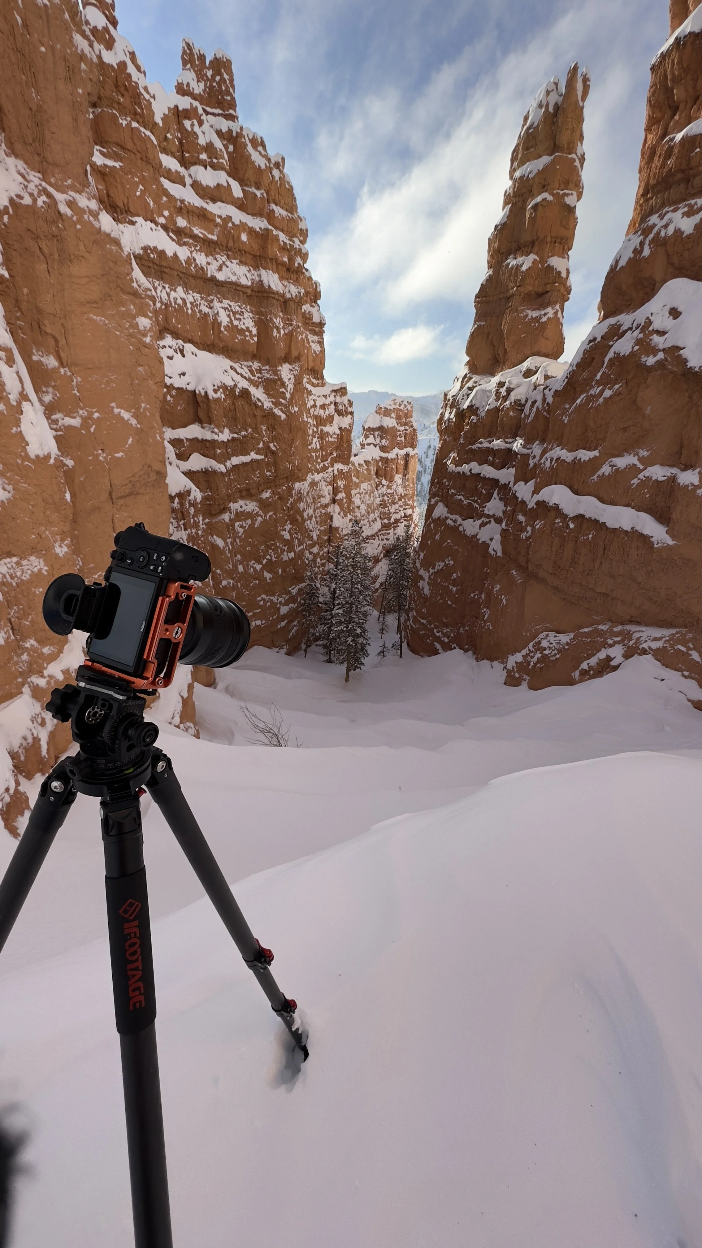 A camera on a tripod set up in a snow-covered canyon with tall orange rock formations and a cloudy blue sky above.