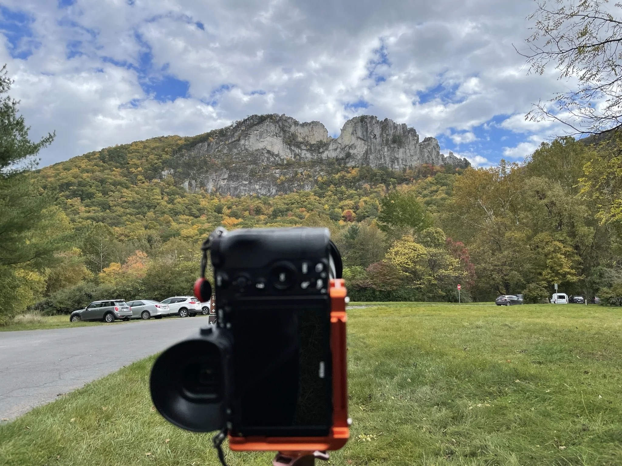 A camera on a tripod captures a scenic view of mountains and a forest with colorful autumn trees, parked cars on a road, and a partly cloudy sky.