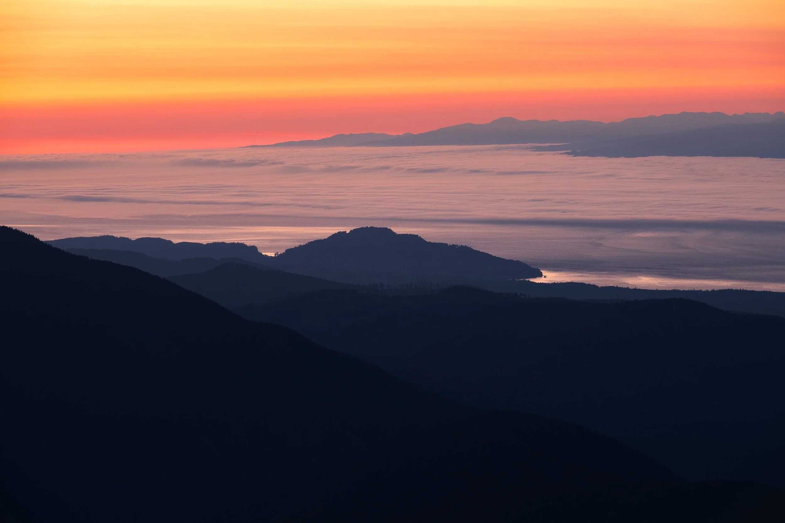 Sunset over the misty landscape of Vancouver Island, Canada, with a colorful horizon illuminating the ocean and silhouetted mountains.
  Coastal, Atmospheric, Golden Hour, Light  