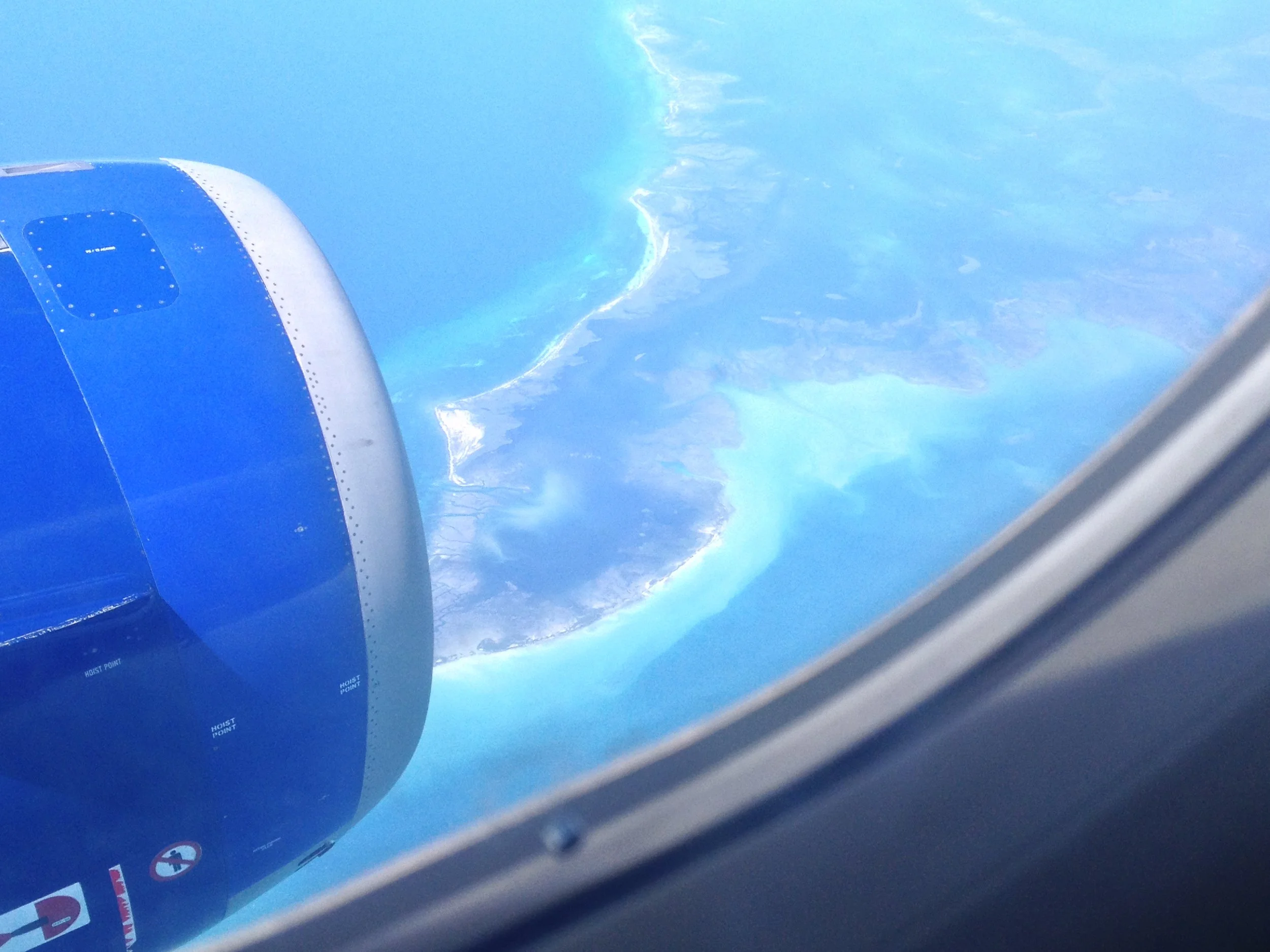A view from an airplane window showing a blue aircraft engine and a coastline with sandy beaches and turquoise water.