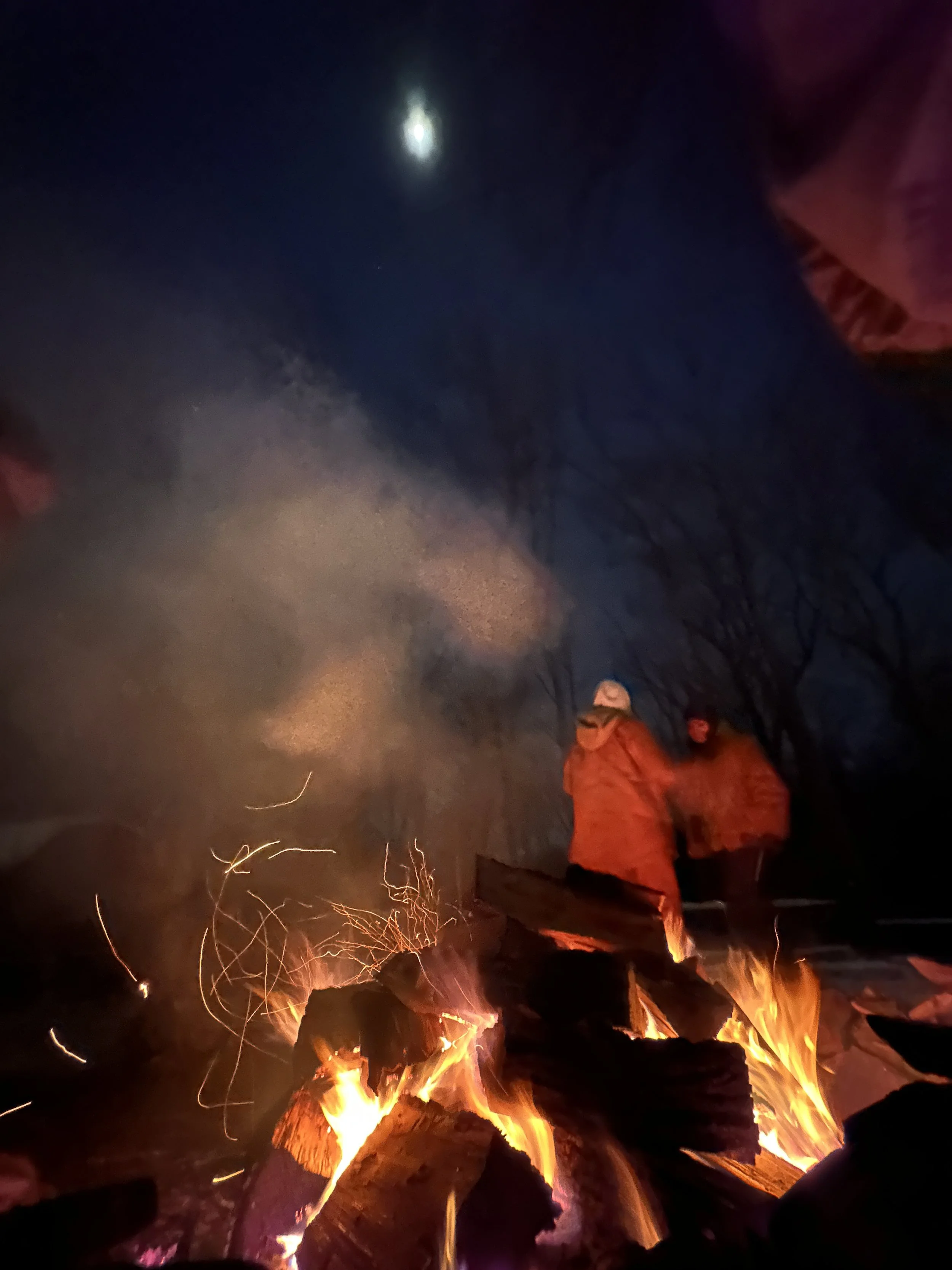 People gathered around a campfire at night with a visible moon and dark sky in the background.