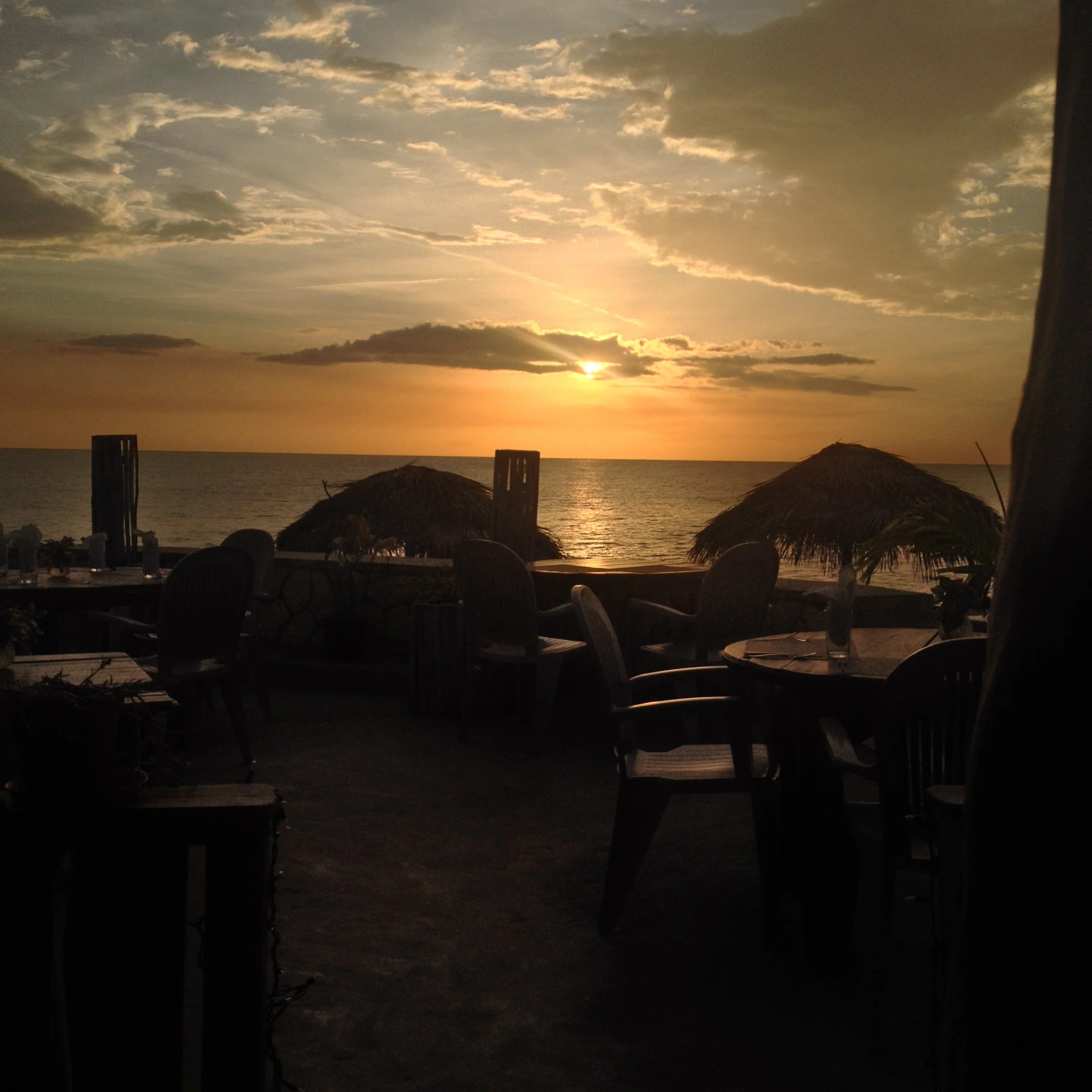 Sunset view over the ocean seen from a beachside restaurant with silhouettes of tables, chairs, and thatched umbrellas in the foreground.