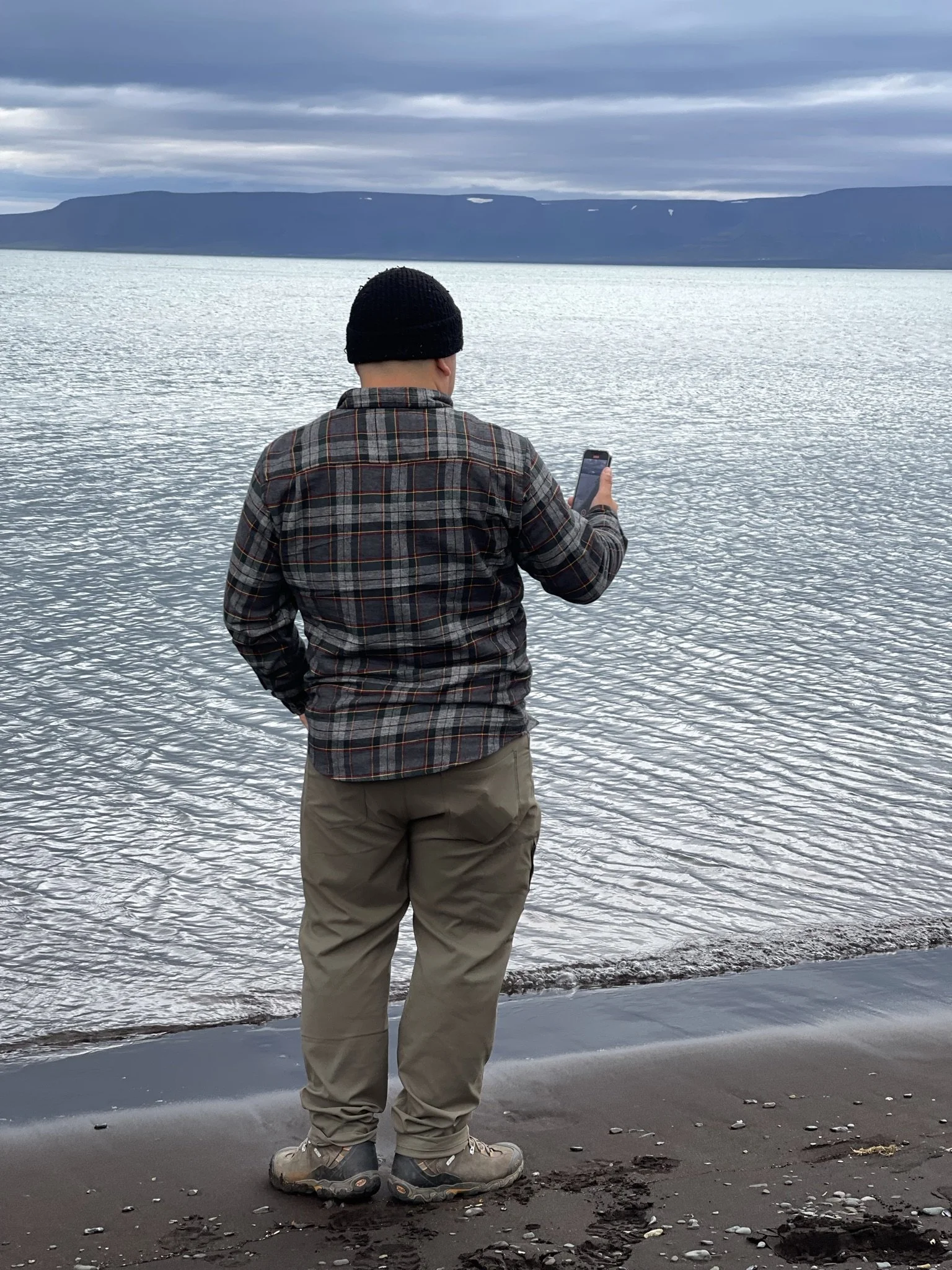 Man in outdoor clothing standing on a sandy beach, looking at a smartphone, with a large body of water and mountains in the background.