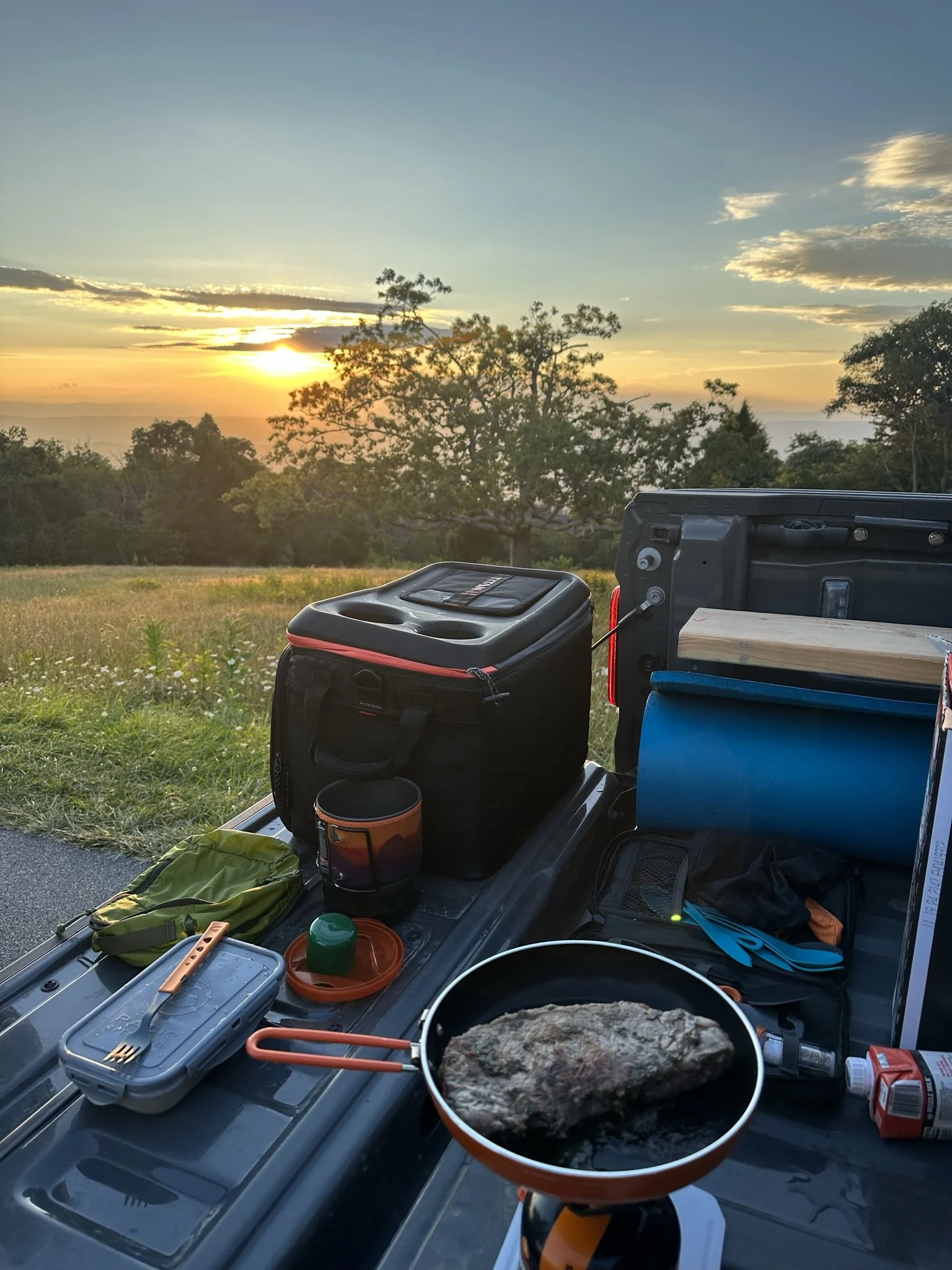 Pickup truck bed with camping gear and food, preparing a meal at sunset in a scenic outdoor location.