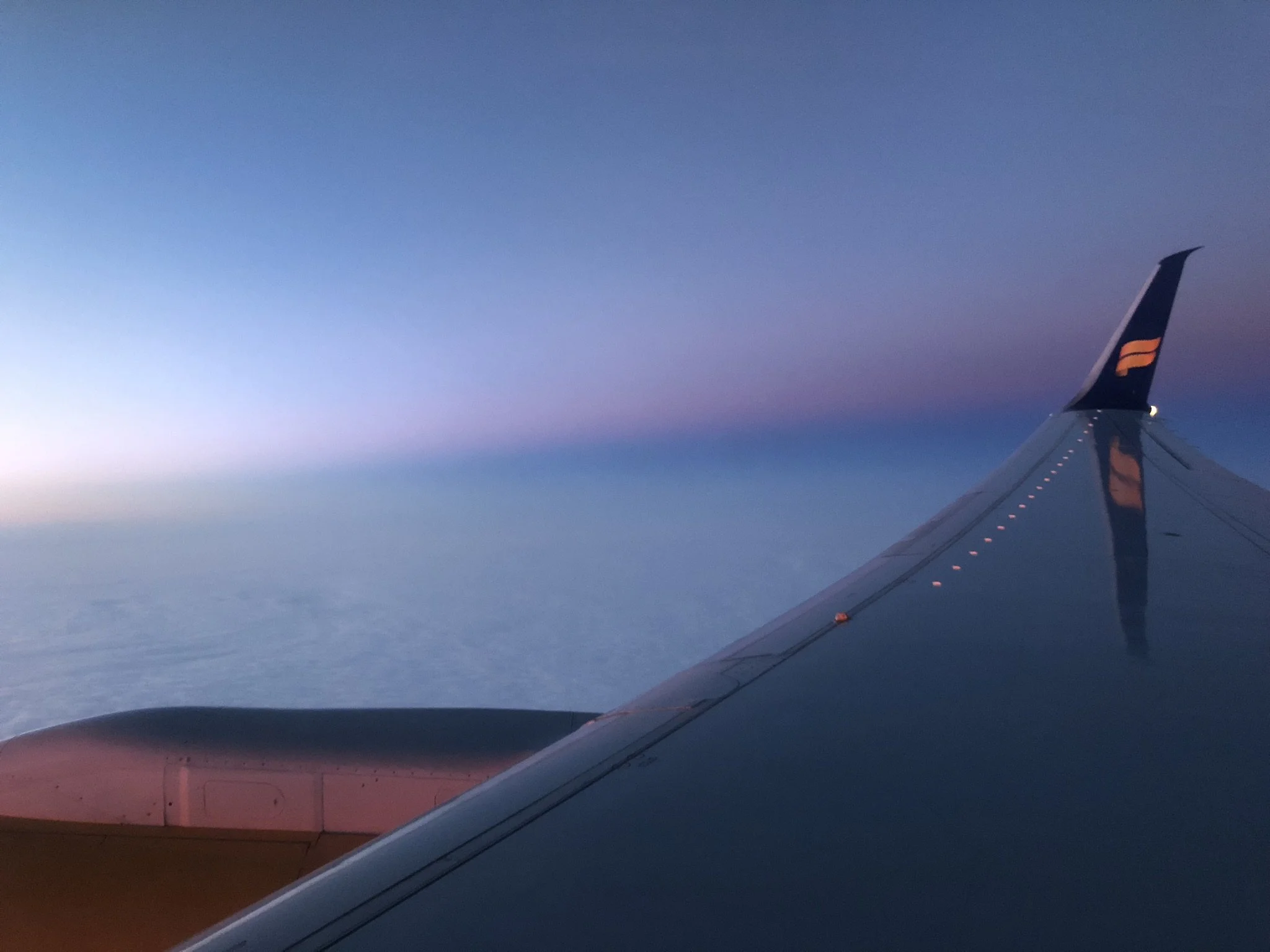 A view from an airplane window showing the wing and the sky during sunset or sunrise with pastel shades of purple, pink, and blue.