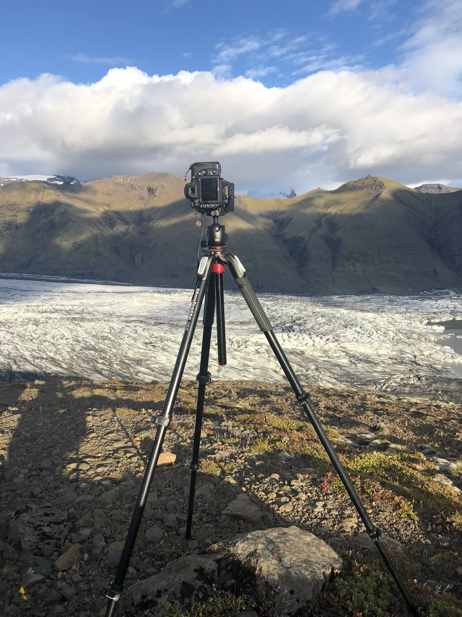 Camera mounted on tripod set up on rocky terrain, overlooking a glacier with mountains in the background under a partly cloudy sky.