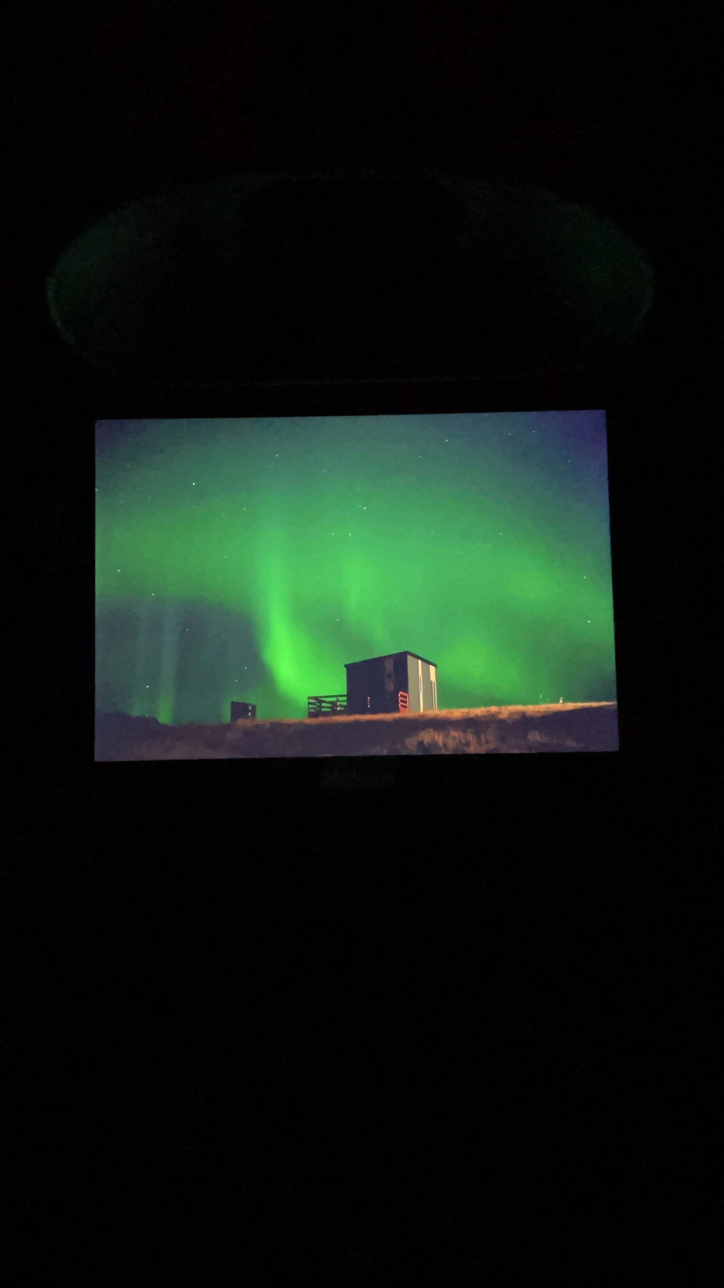 Photo of a phone screen showing the Northern Lights over a small house or cabin at night.