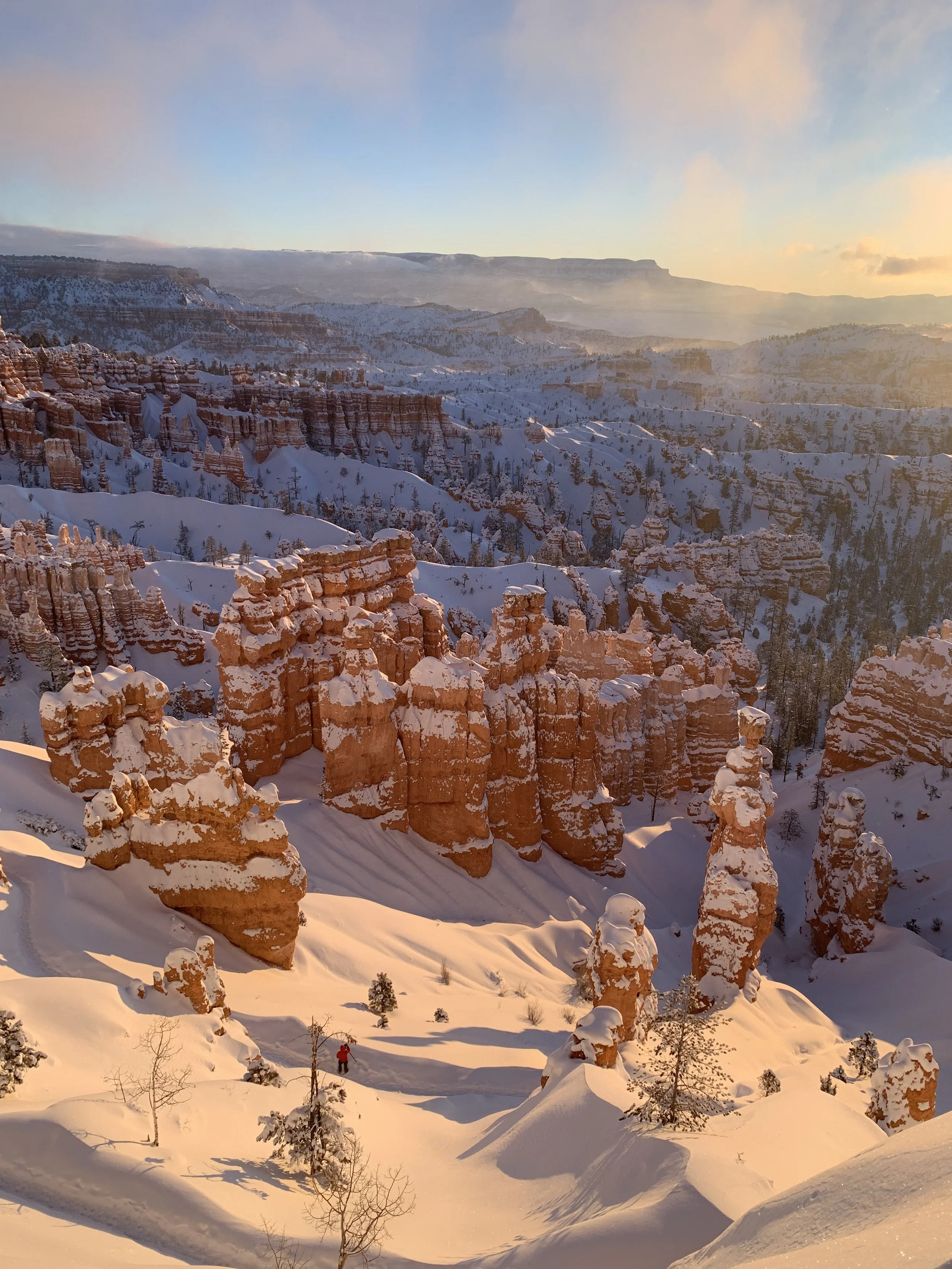 Snow-covered canyon with tall, orange rock formations and a lone person in red clothing walking through the snow at sunset.