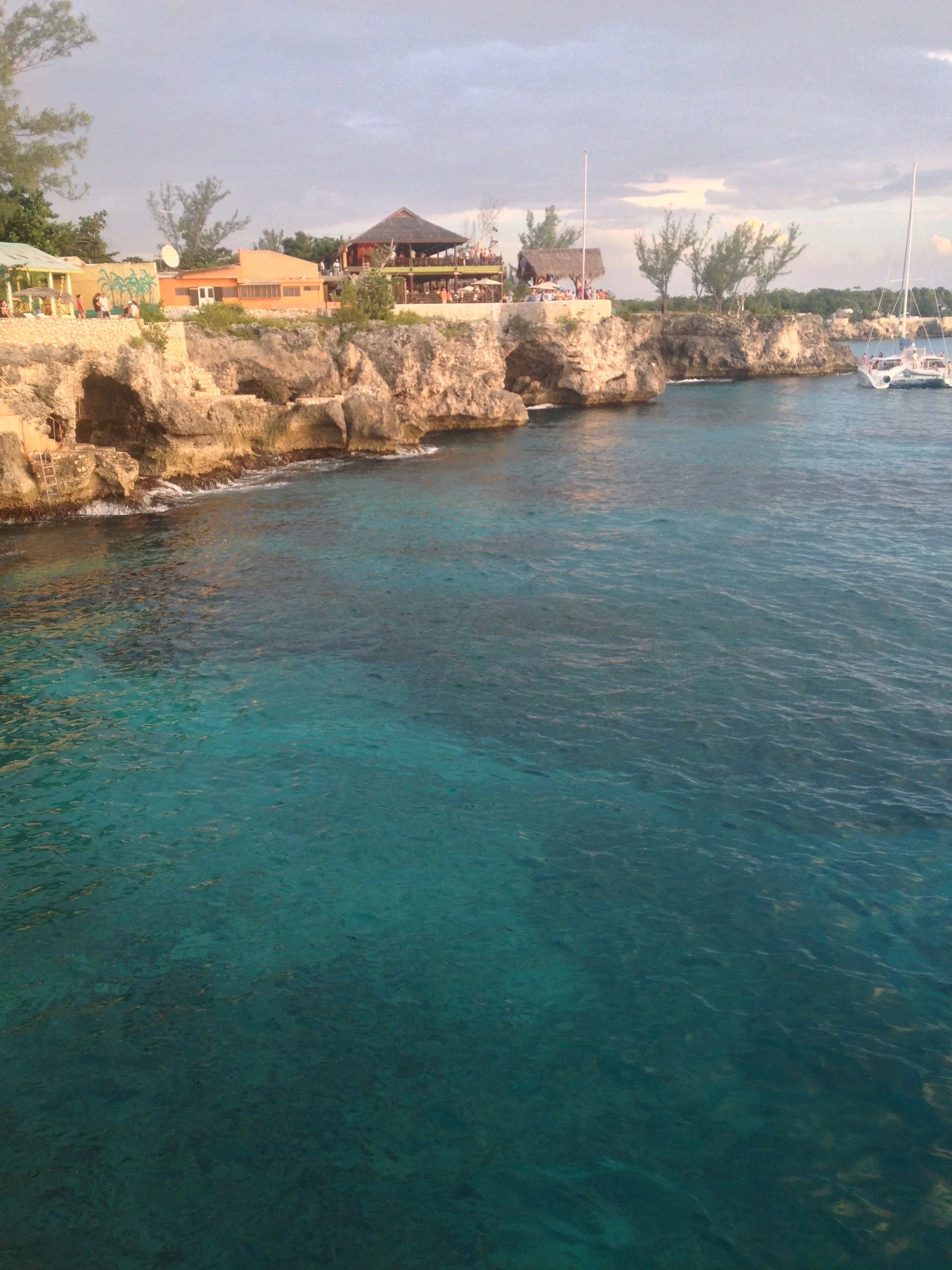A seaside view with clear, turquoise water in the foreground, rocky cliffs along the shoreline, and a coastal restaurant with outdoor seating on the cliff top. Several sailboats are anchored in the water, and there are trees and a cloudy sky in the b