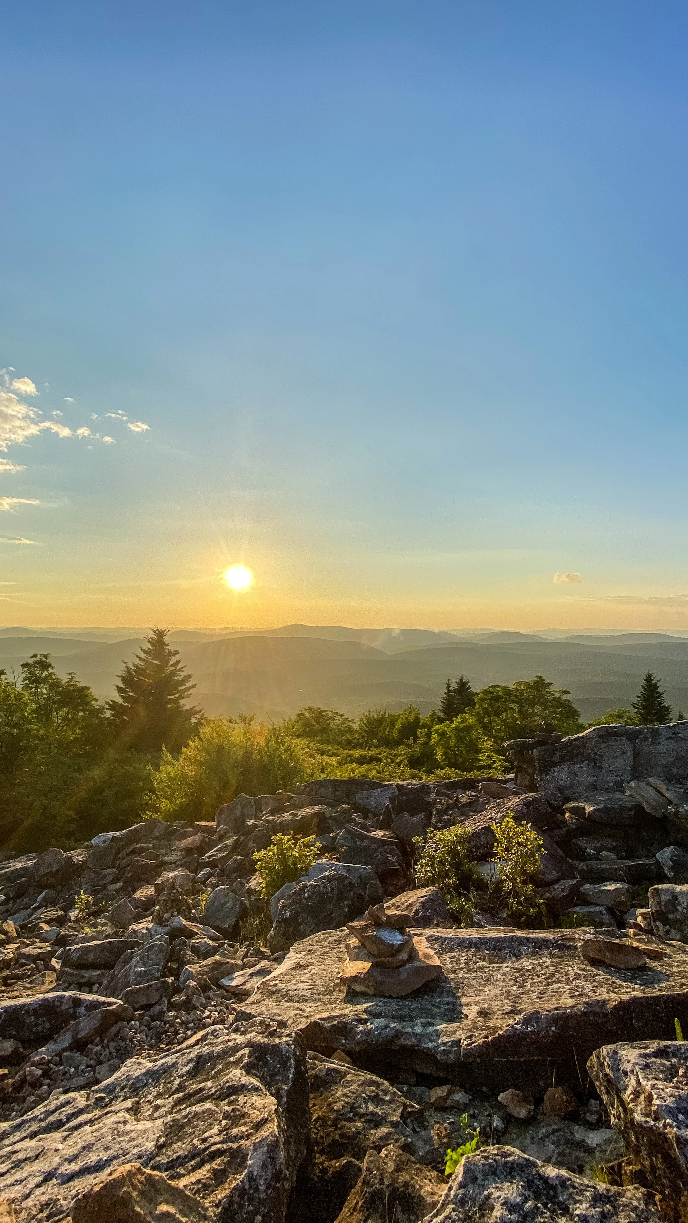 Sunset over a rocky mountain landscape with green trees and layered mountain ranges in the background.