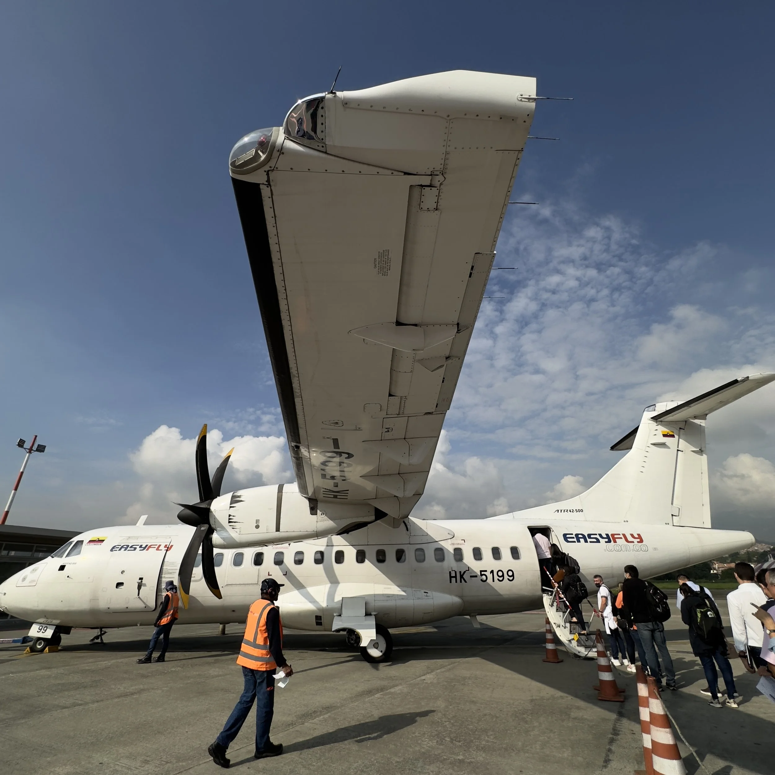 Passengers boarding an EasyFly regional turboprop airplane at the airport, with ground crew and orange safety cones present.