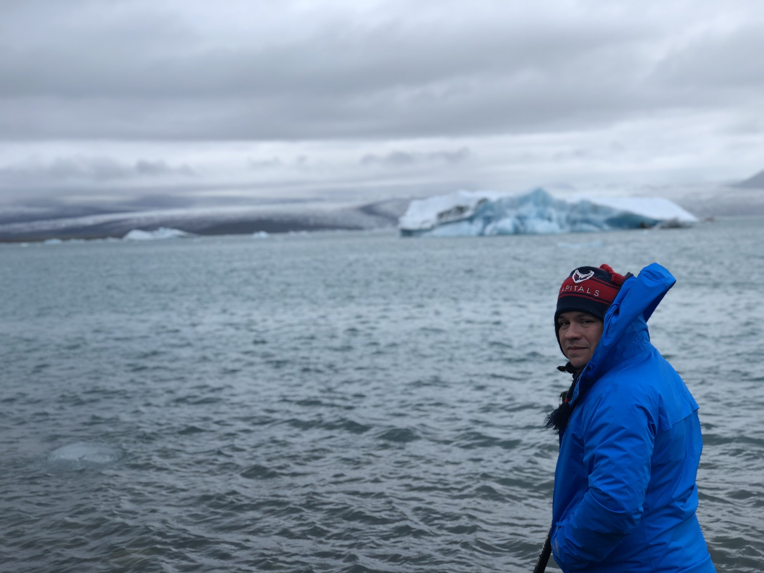 A man in a blue jacket and a red and blue hat standing near water with icebergs and snow-covered land in the background under cloudy skies.