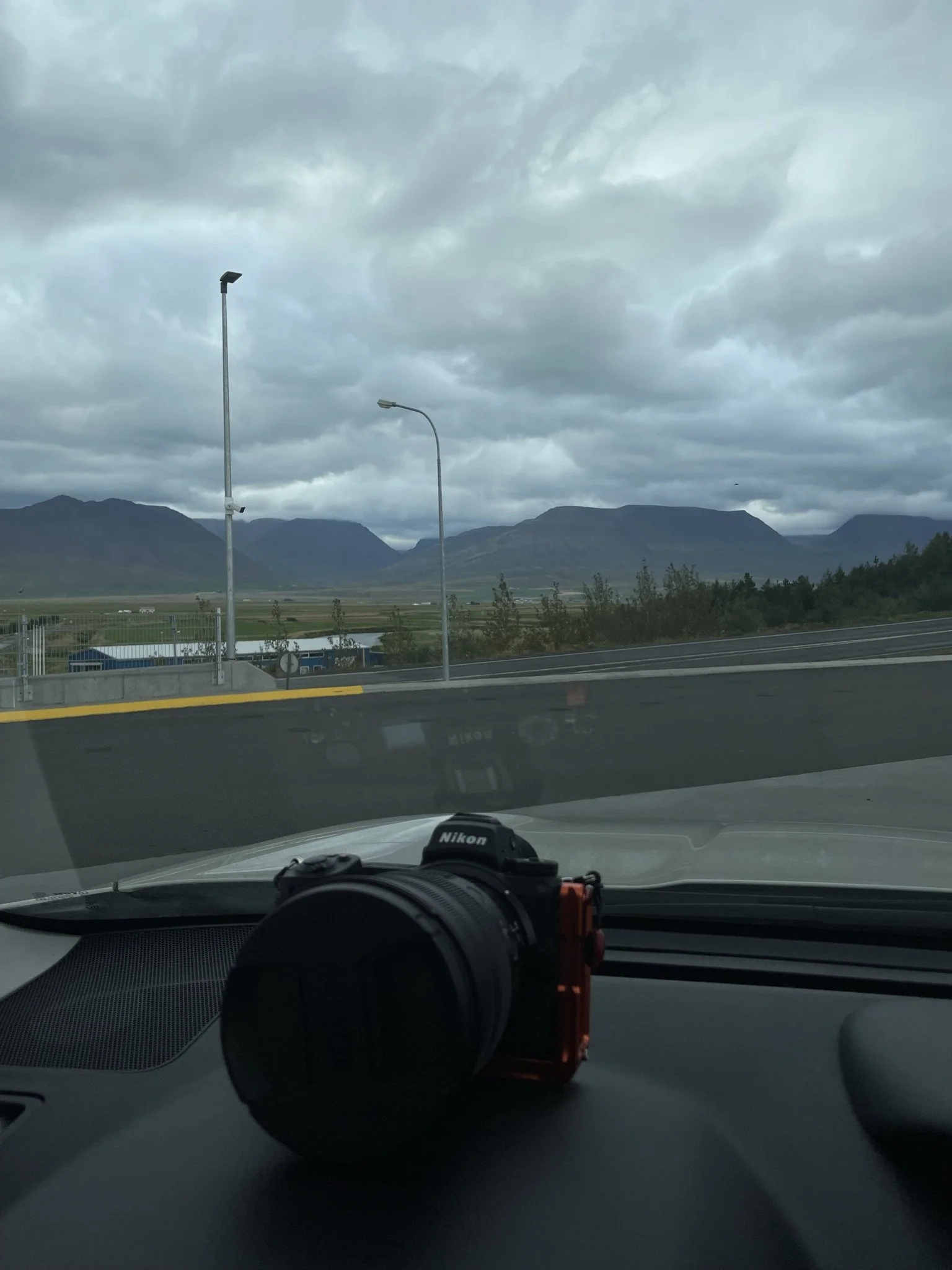 View from inside a car showing a Nikon camera on the dashboard, with a cloudy sky, mountains, and a strip of road in the background.