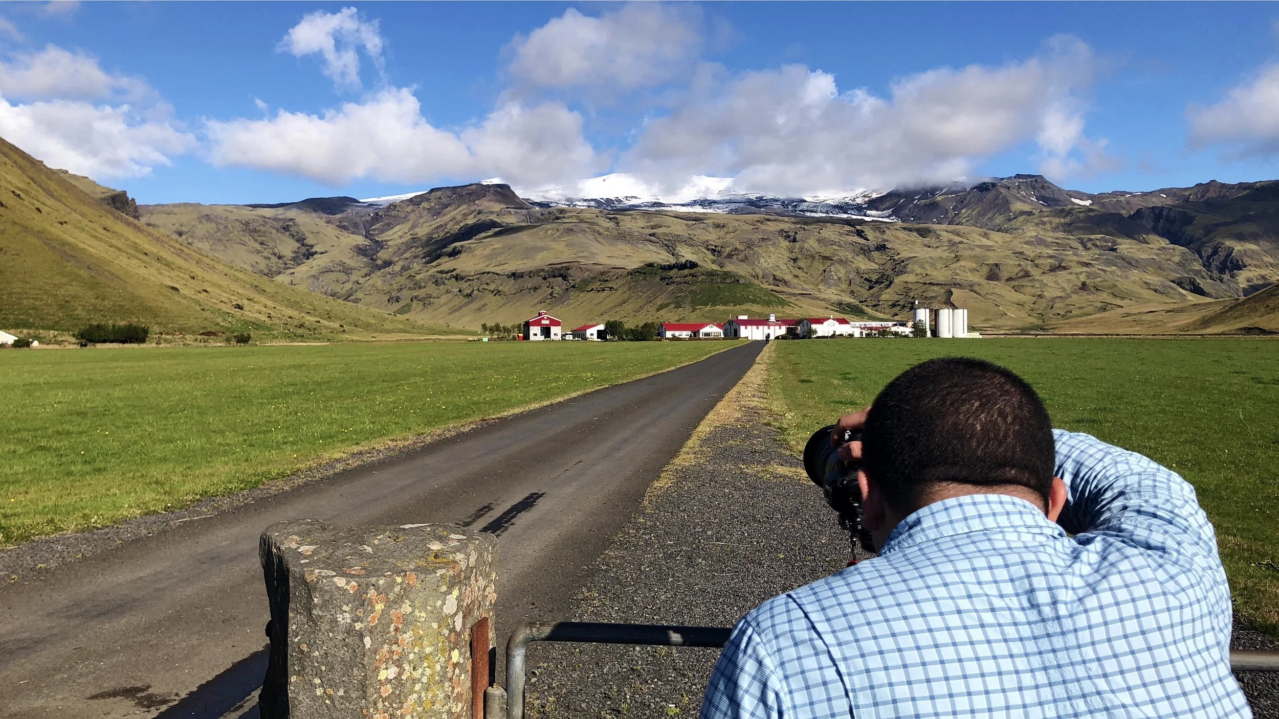 A man in a blue checkered shirt taking a photograph of a farm with white buildings and red roofs, surrounded by green fields and mountains in the background under a partly cloudy sky.