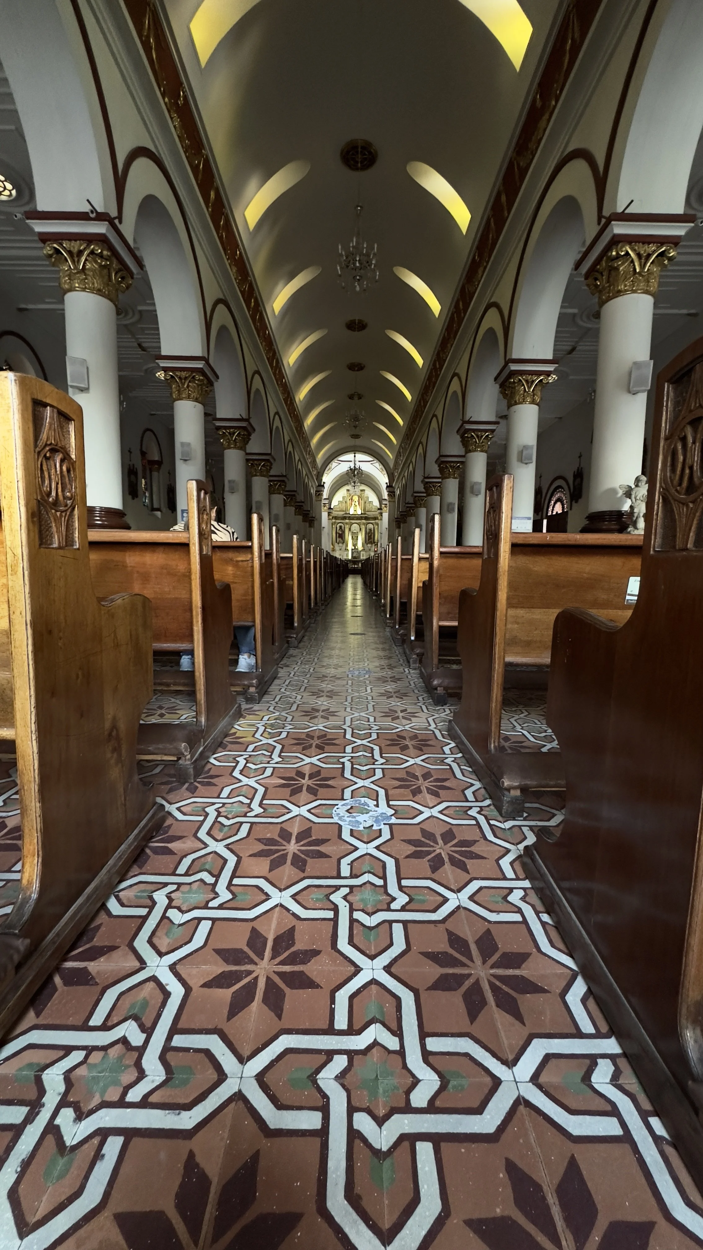Interior of a church or cathedral with wooden pews, ornate columns with gold accents, and a decorated tiled floor leading to an altar at the front.