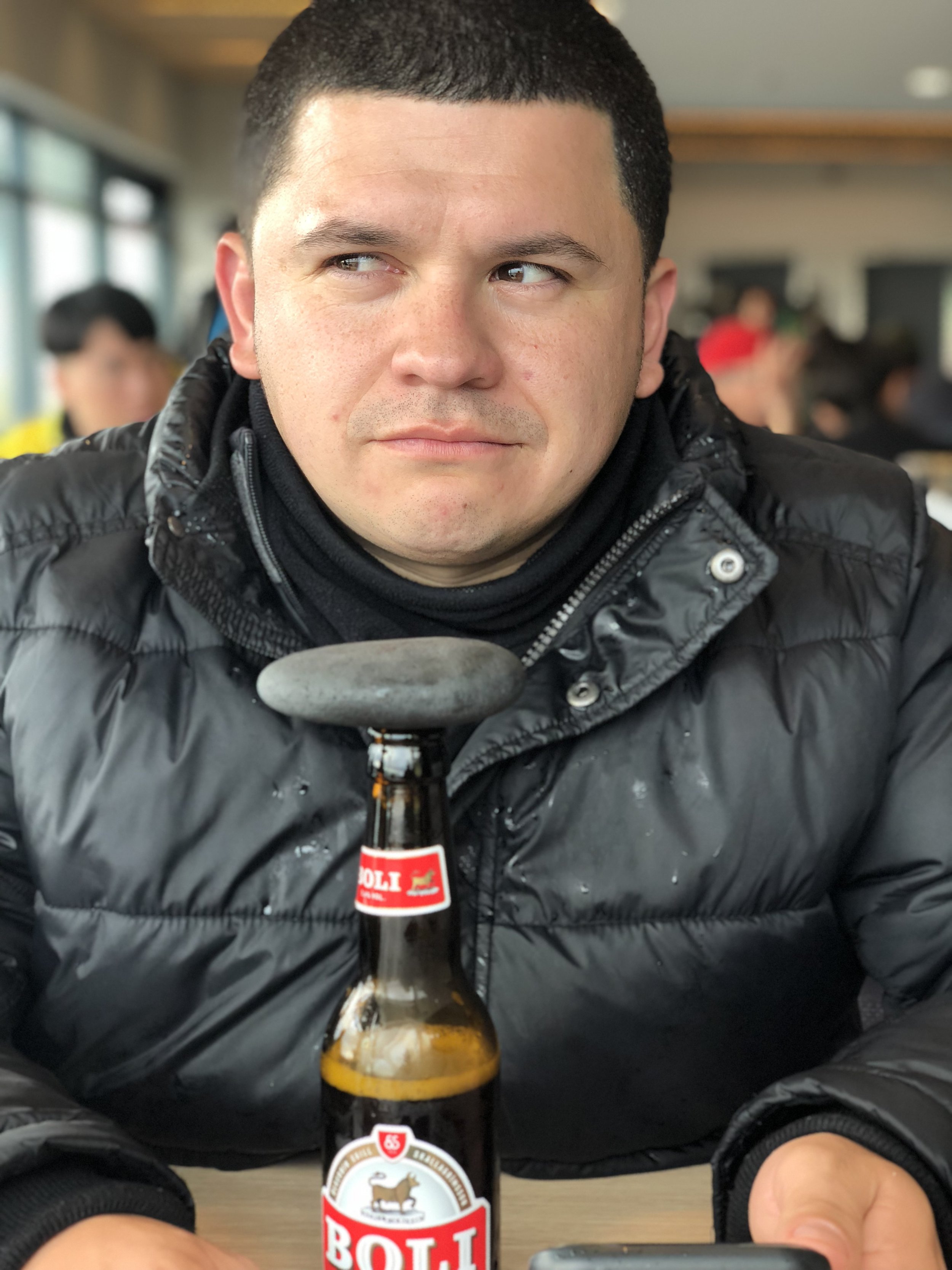 A young man with short dark hair and a fair complexion sitting indoors at a table with a bottle of beer in front of him. He is wearing a black puffer jacket and has a neutral expression, looking slightly to the left. The background shows other people
