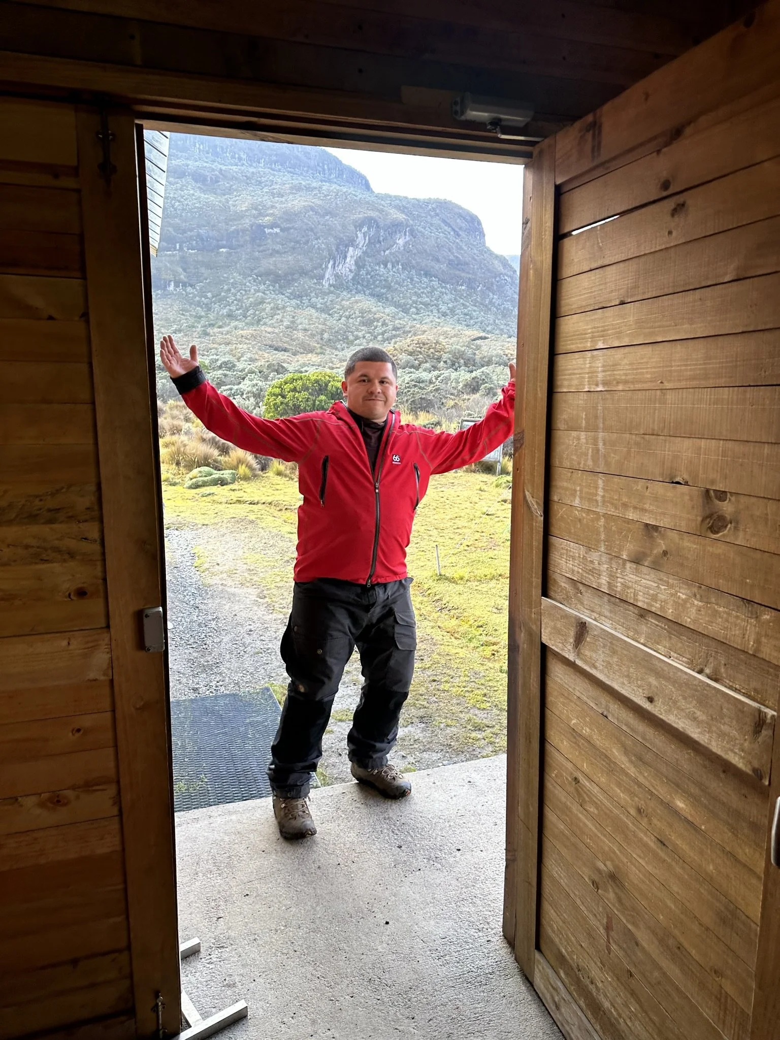 A man in a red jacket and black pants standing outside holding open a wooden door with a mountain landscape in the background.