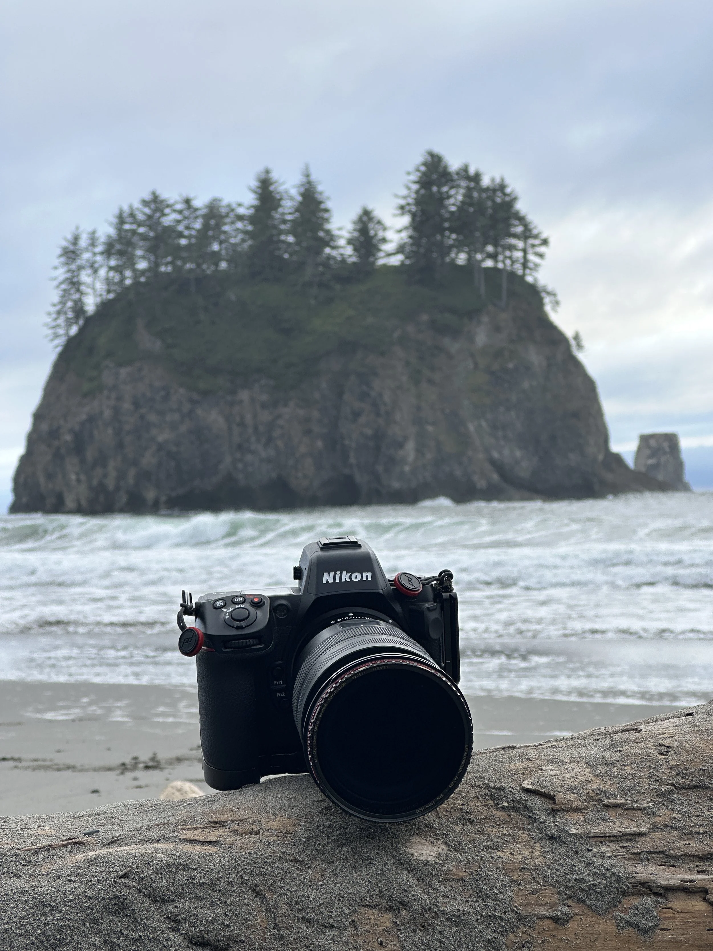A Nikon camera on a rock at the beach with an island with trees in the background and an overcast sky.