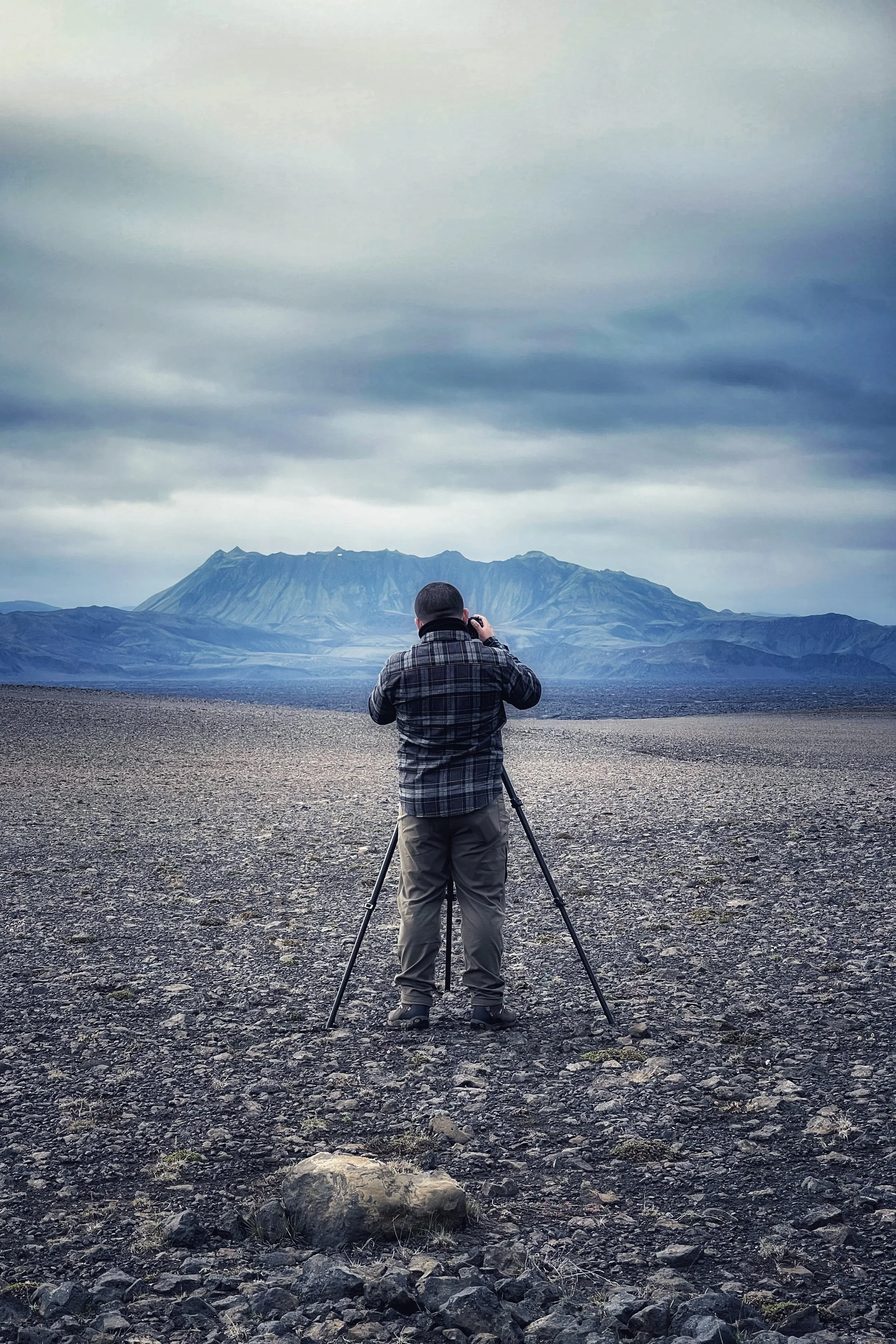 A person standing on rocky terrain, taking a photograph of a mountain in the distance, under a cloudy sky.