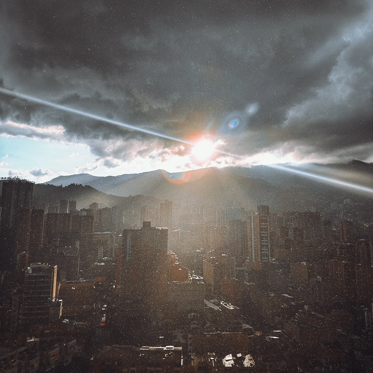 City skyline with tall buildings, mountain range in the background, and a dramatic sky with dark clouds and the sun partly obscured, creating bright sunbeams and lens flares.
