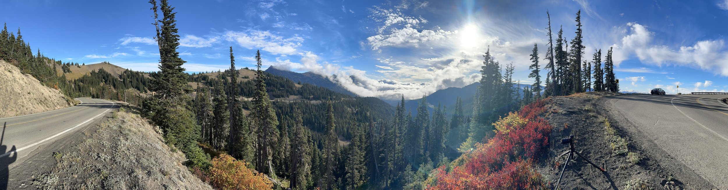Scenic mountain road winding through a forested landscape with tall evergreen trees, colorful autumn foliage, and a car parked on the side, with mountains and a cloudy sky in the background.