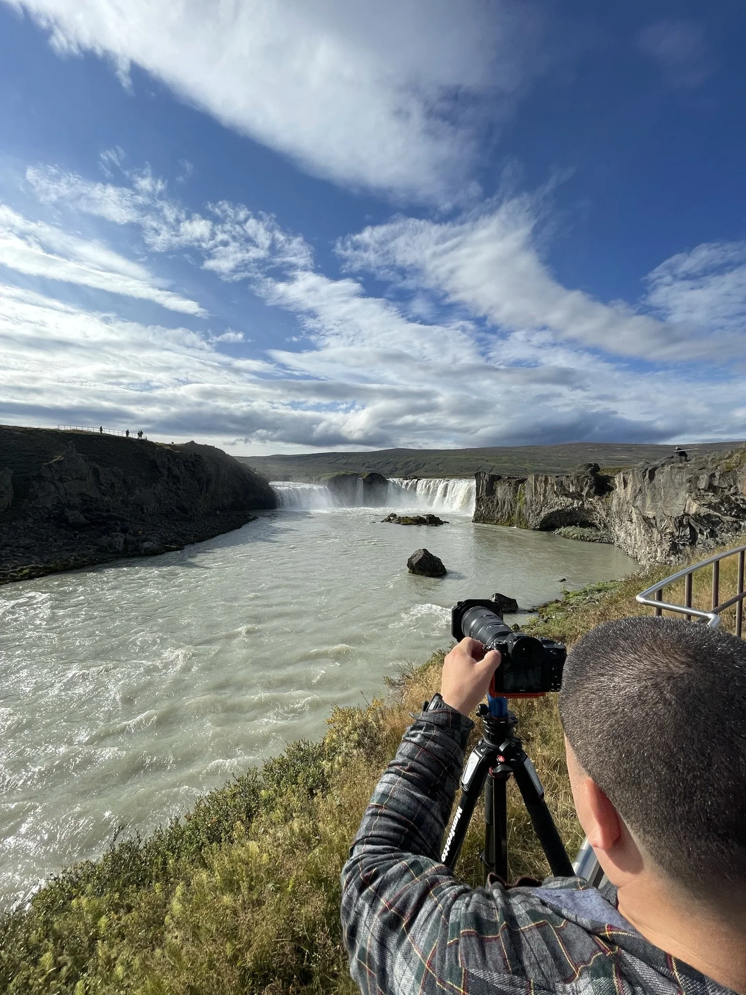 A person with short hair taking a photo of a waterfall with a camera on a tripod, beside a railing on a grassy area, under a partly cloudy sky.