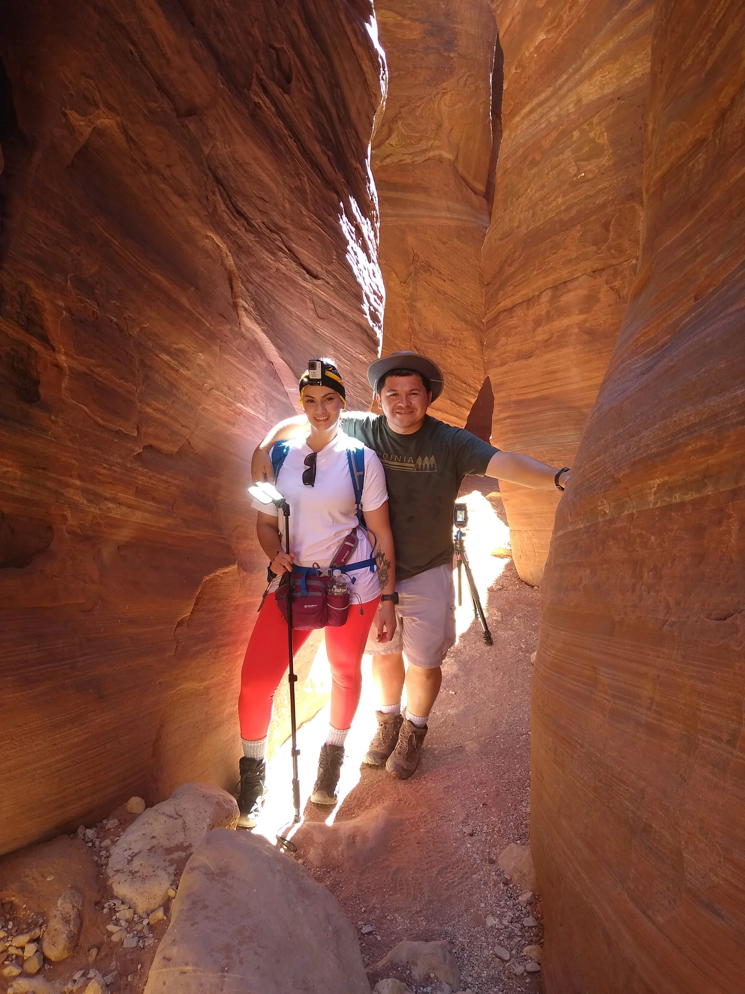 Two hikers standing in a narrow slot canyon with orange and red layered rock walls, smiling at the camera, capturing a moment of adventure.