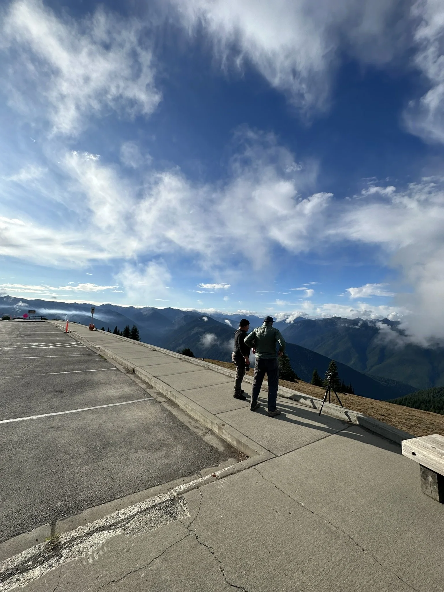 Two people standing on a sidewalk overlooking mountain ranges, with a camera set up on a tripod nearby, under partly cloudy sky.