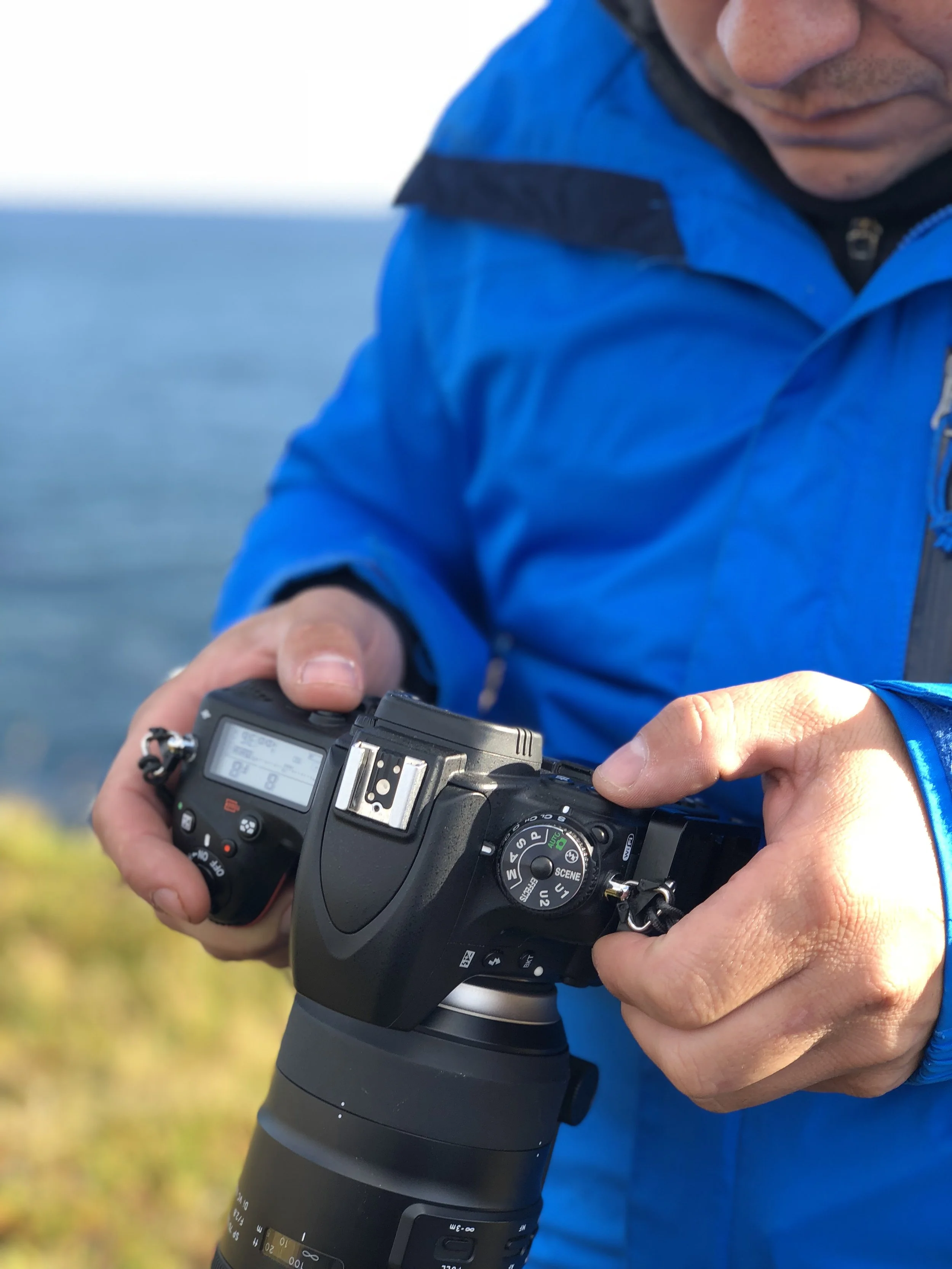 A person in a blue jacket holds a DSLR camera outdoors near water, adjusting settings.