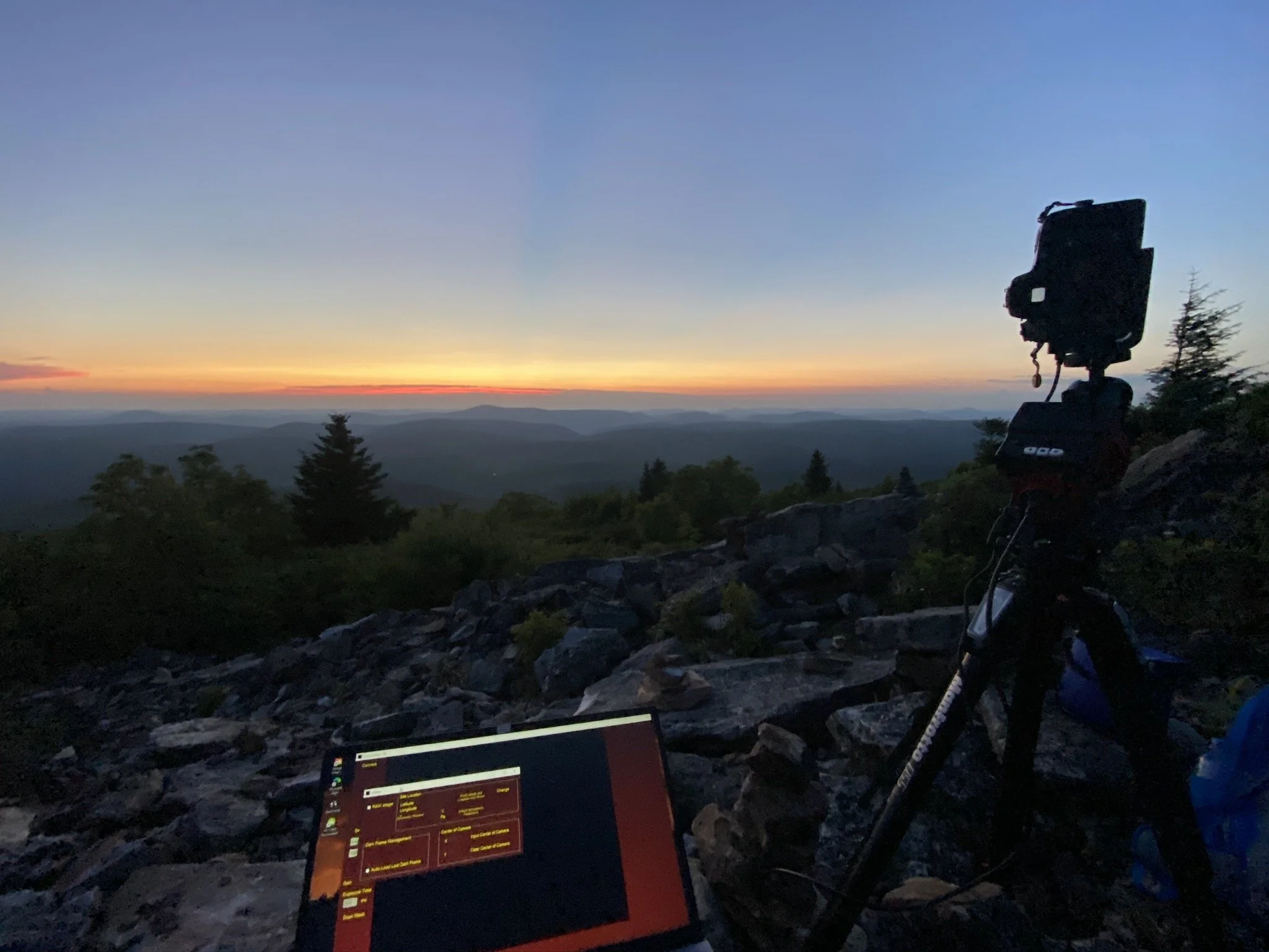 Camera on a tripod and a laptop on rocky terrain overlooking a sunset over a mountain range with trees in the foreground.