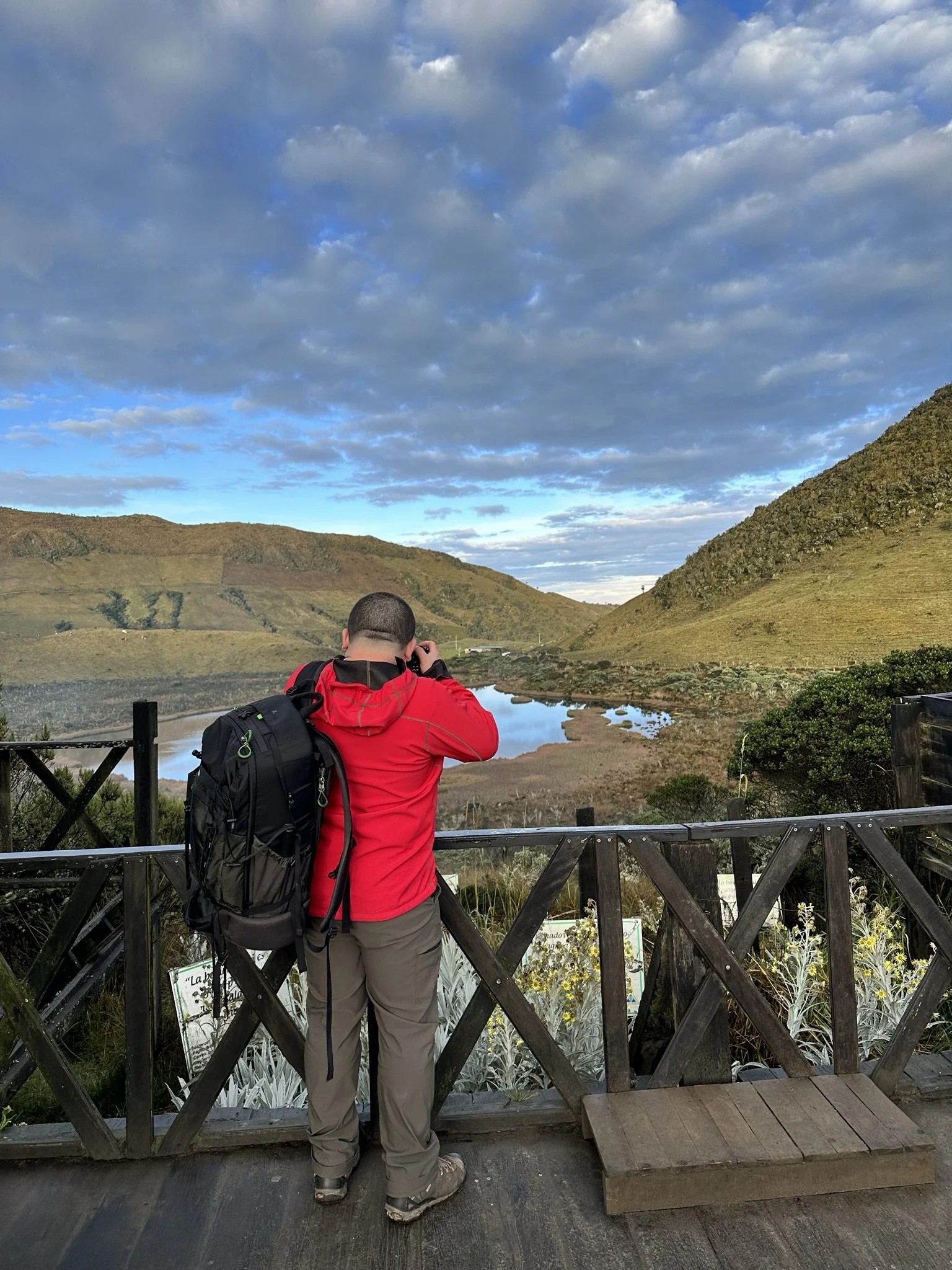 A man in a red jacket and khaki pants with a backpack takes photographs of a scenic landscape with hills, a river, and a cloudy sky from a wooden observation deck.