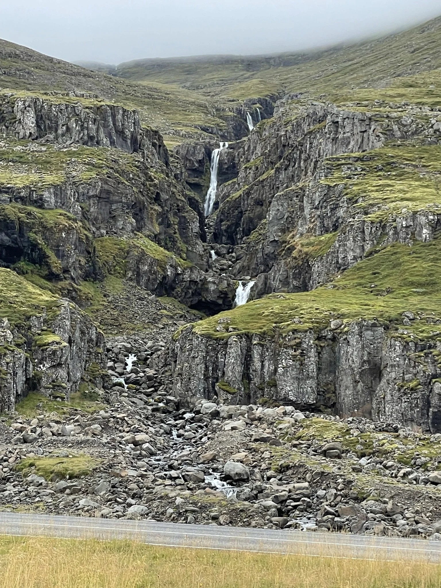 A rugged mountain landscape with waterfalls cascading down rocky cliffs and green moss-covered slopes under a foggy sky.