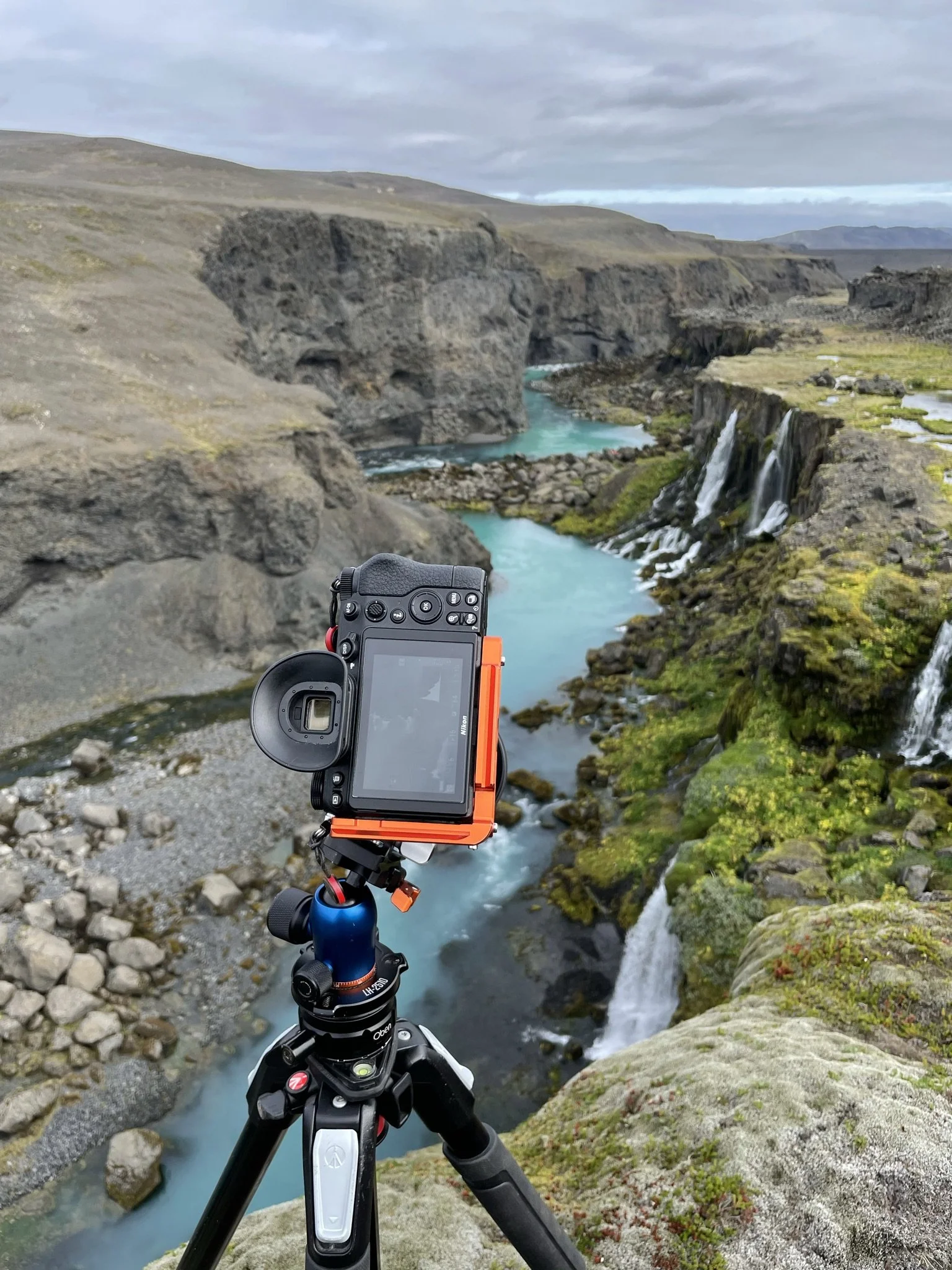 A camera on a tripod capturing a landscape of a river with waterfalls, moss-covered rocks, and rugged cliffs under a cloudy sky.