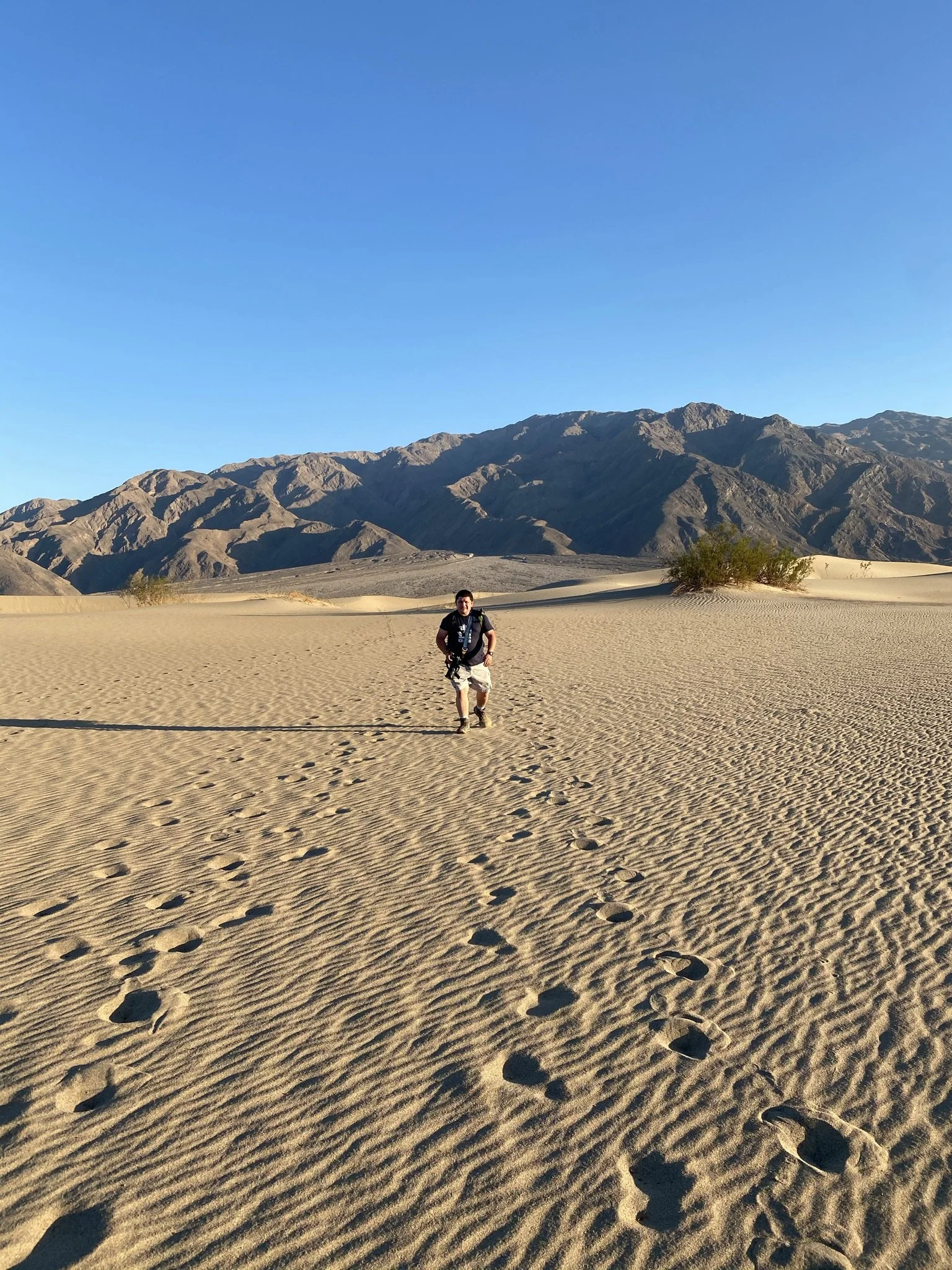 A man walking across sand dunes in a desert with mountains in the background and clear blue sky overhead.