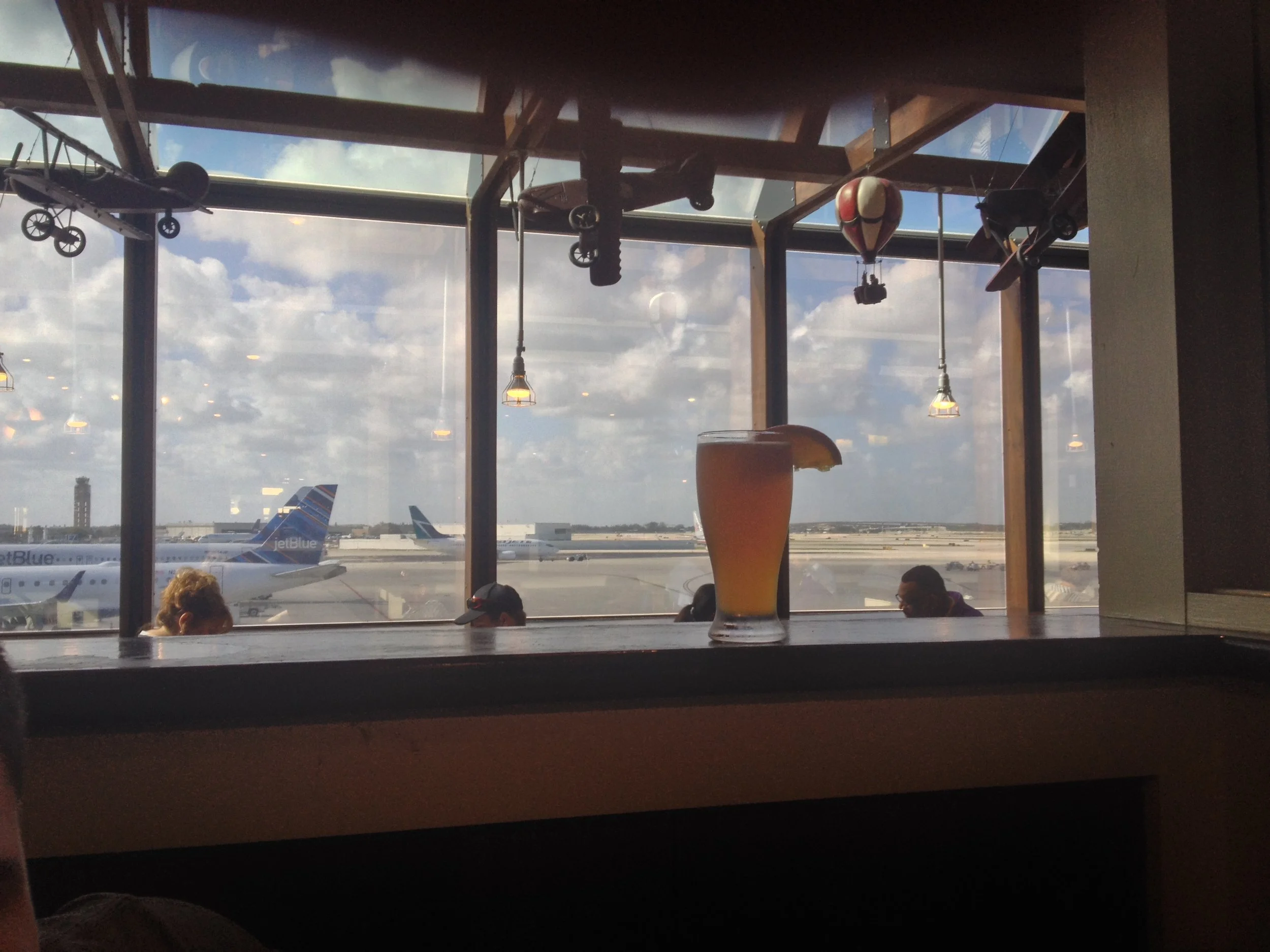 A glass of orange-colored beverage with a lemon wedge on an airport lounge table, with airplanes visible through large windows in the background.
