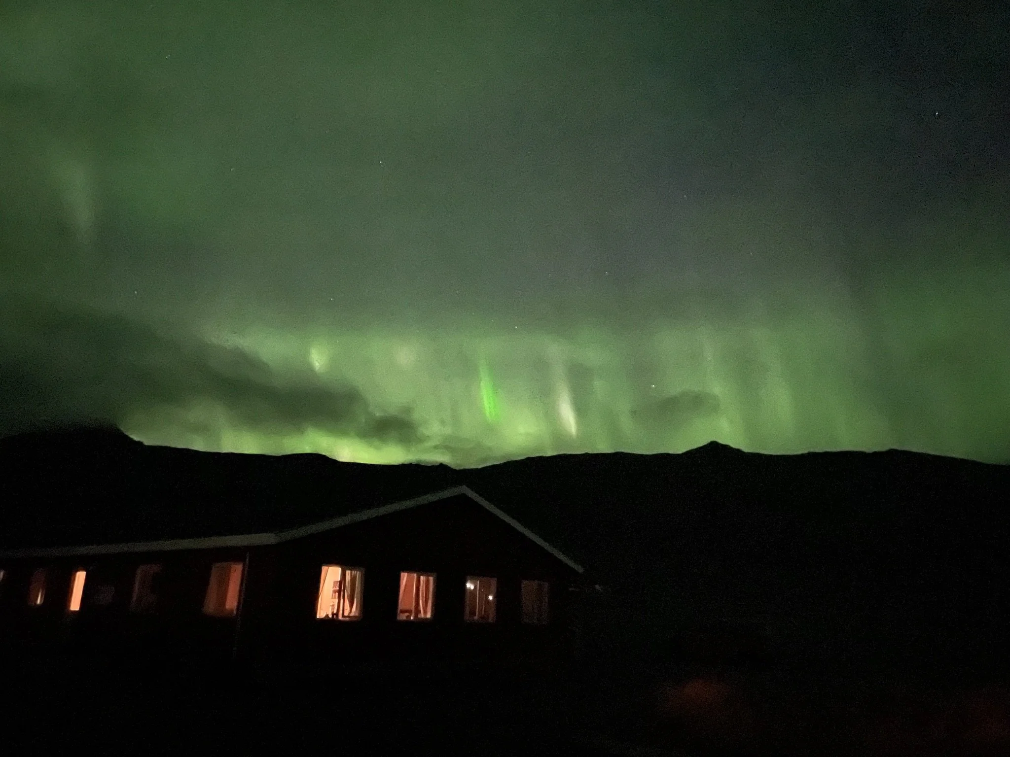 Northern lights illuminating the night sky over a house with lit windows.