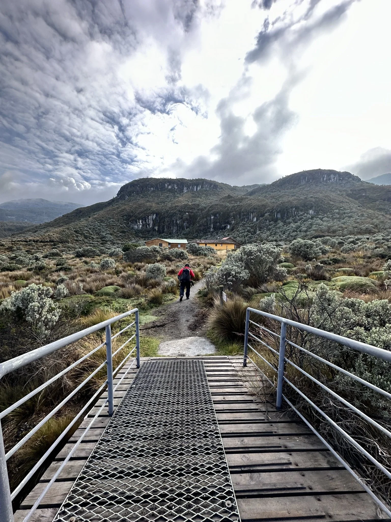 A hiker walking along a trail in a mountainous landscape with shrubs and cloudy sky.