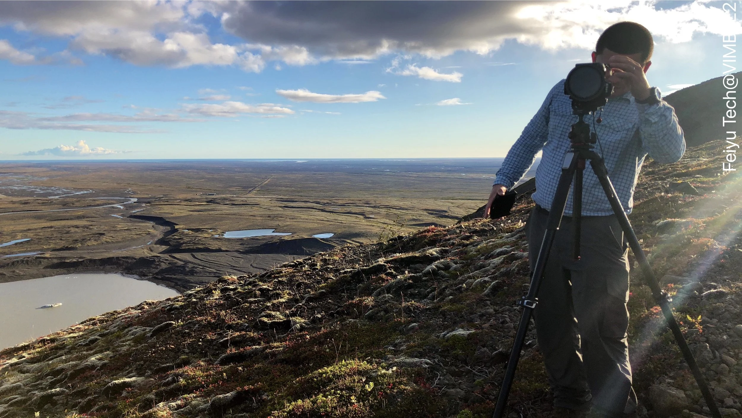 A person standing on a grassy hill taking a photograph with a camera on a tripod, overlooking a vast landscape with rivers and lakes under a partly cloudy sky.