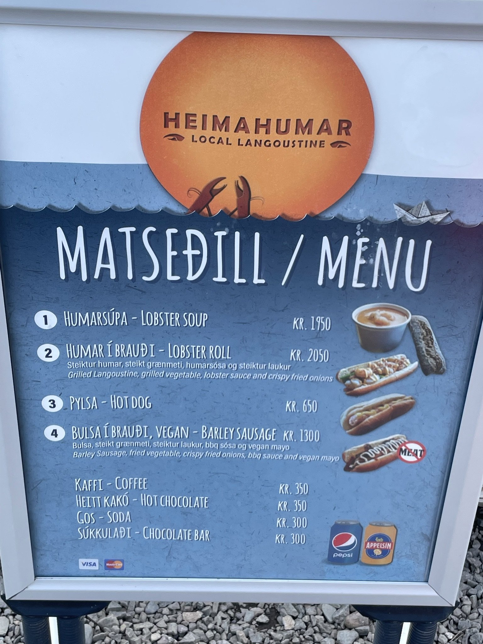 Restaurant menu in Icelandic featuring lobster soup, lobster roll, hot dog, vegan barley sausage, coffee, hot chocolate, soda, and chocolate bar, with images of lobster dish, hot dog, and drinks.