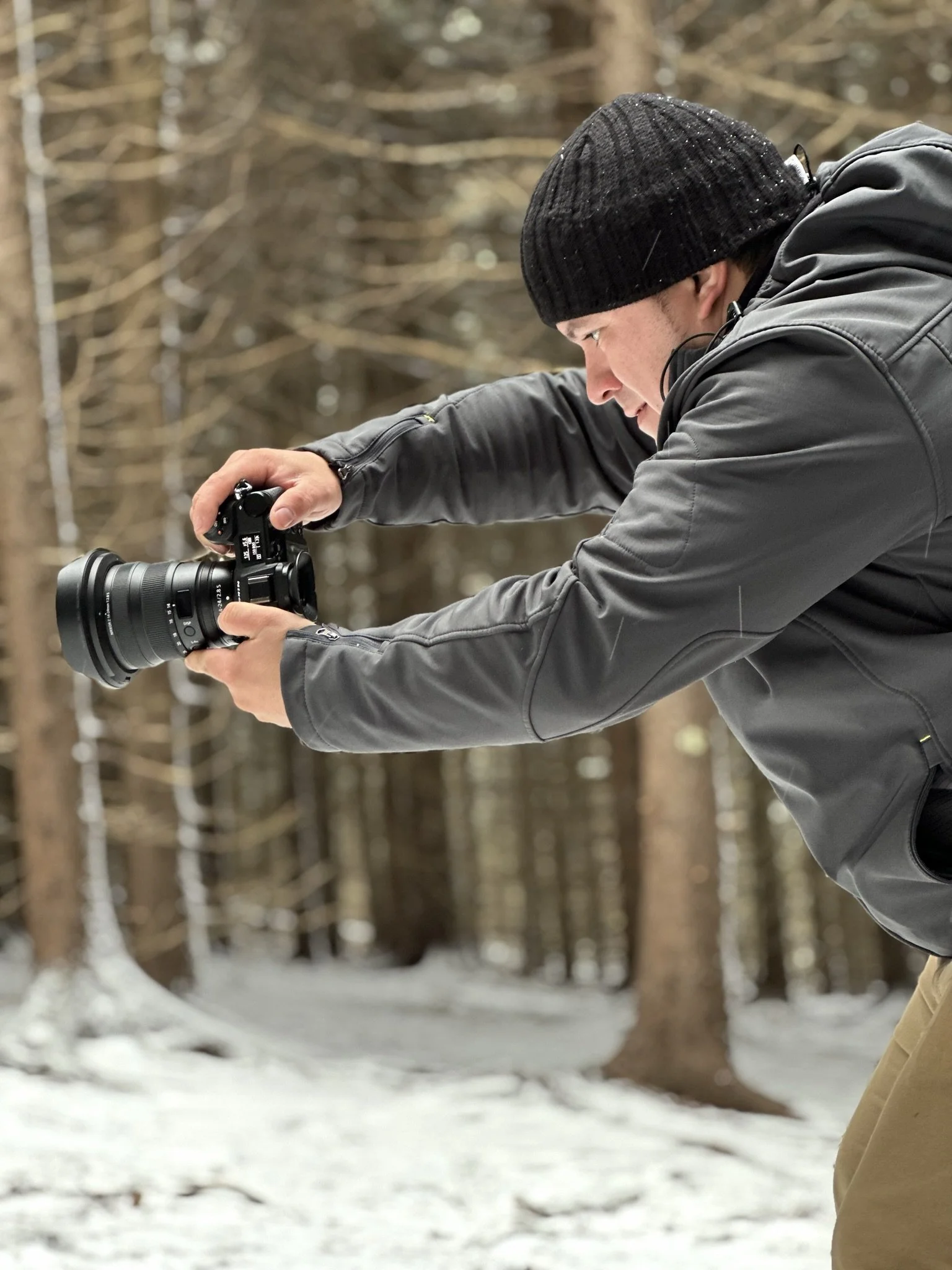 A man in a black beanie and gray jacket taking a photograph with a camera in a snowy forest.