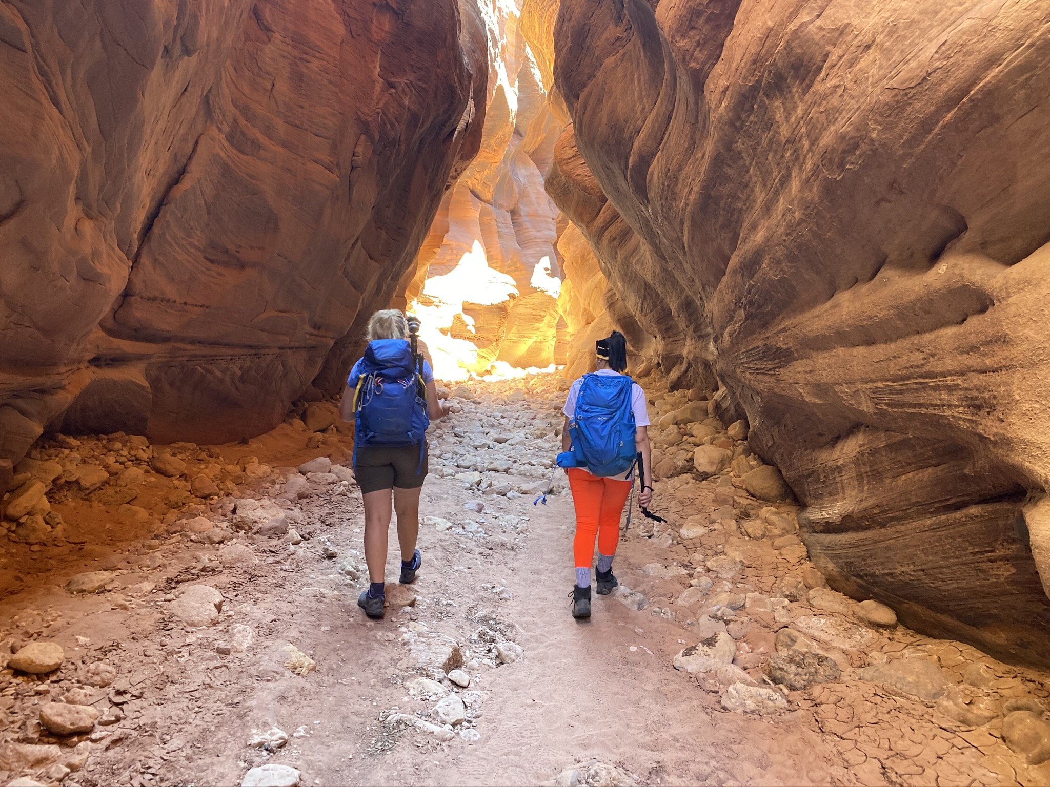 Two women hiking through a narrow, rocky canyon with high, smooth, reddish-orange walls, carrying backpacks.