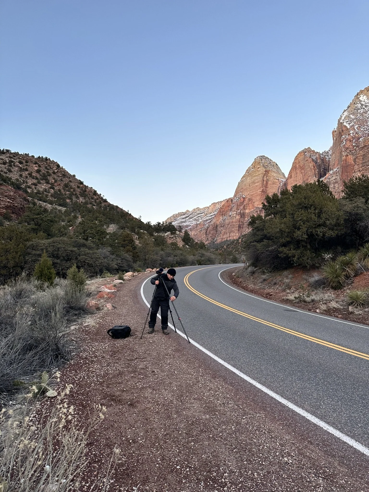 A photographer in black clothing using a tripod on a winding mountain road surrounded by desert plants and red rock formations under a clear blue sky.