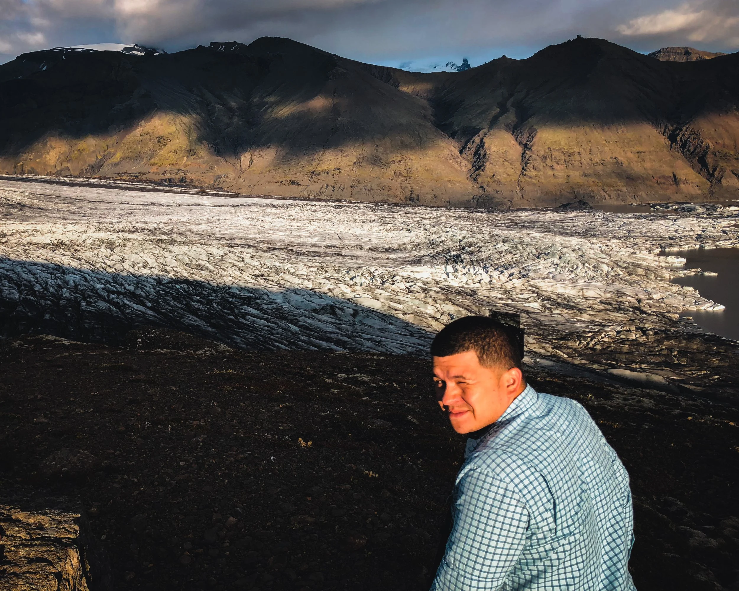 A man wearing a blue checkered shirt winking at the camera with a glacier and mountains in the background.