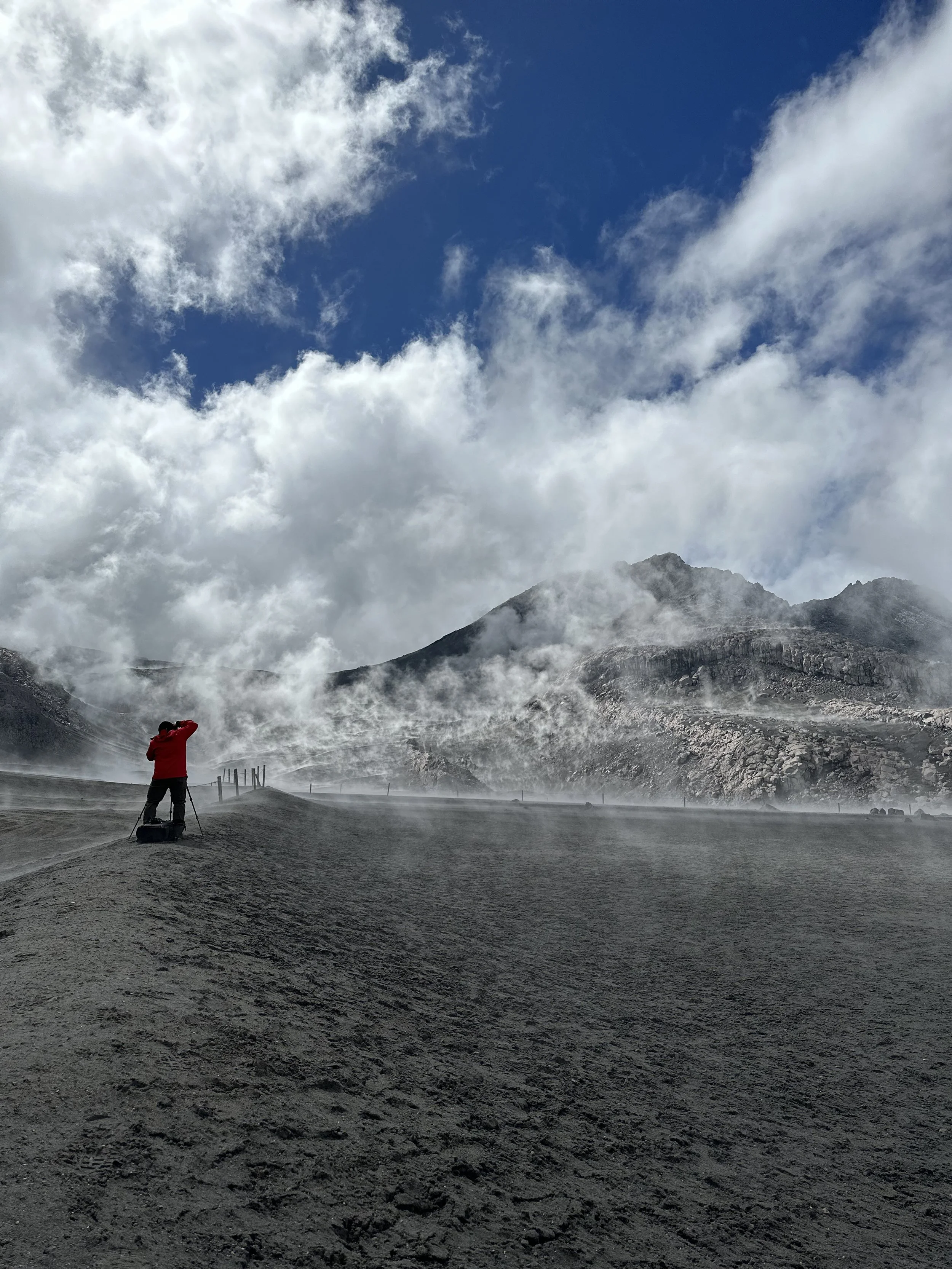 Melser in a red jacket taking photos of a volcanic landscape with steam rising from the ground and mountains in the background under a partly cloudy sky.