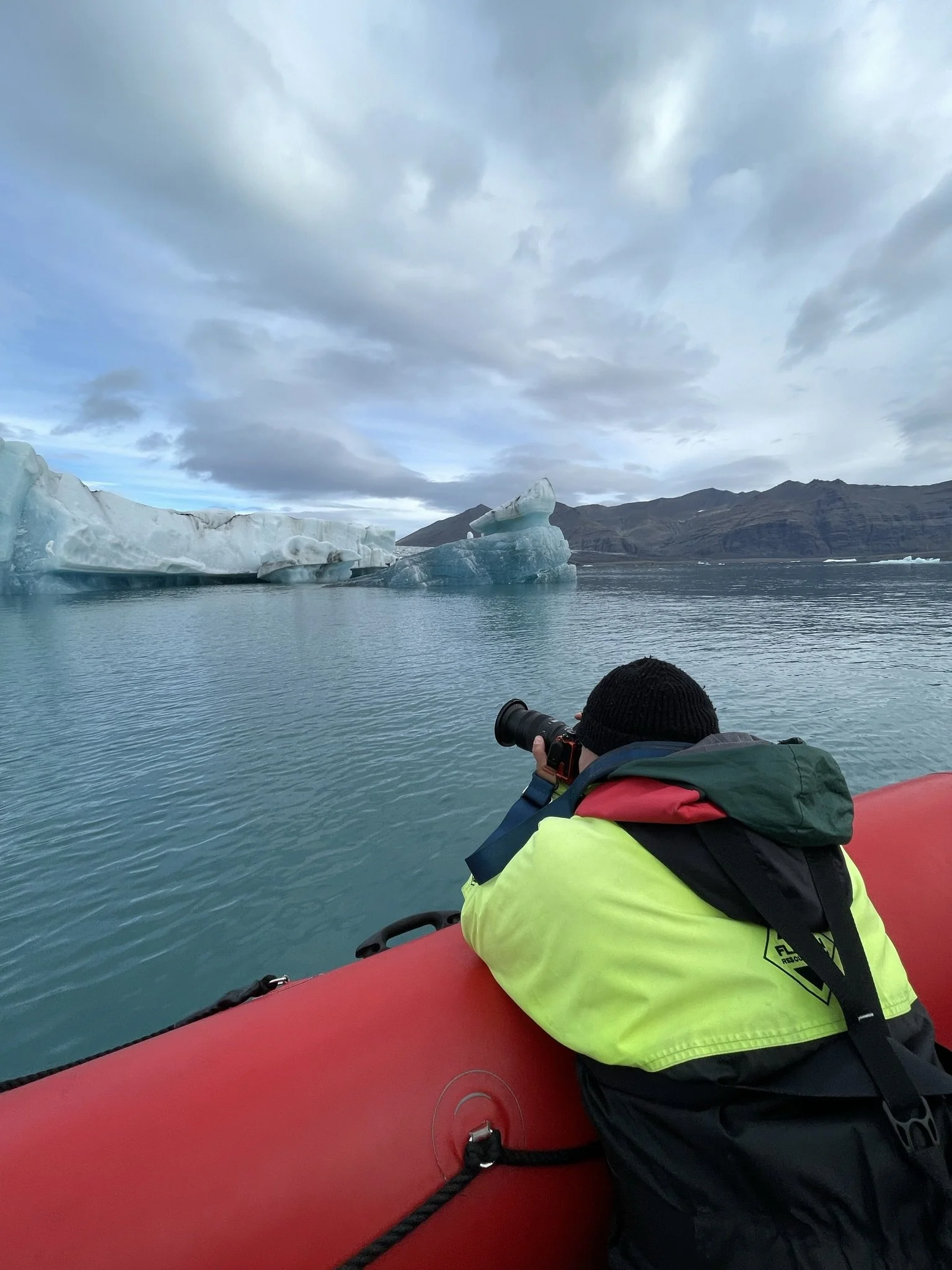 A person in a yellow and black jacket lying on a red inflatable boat, taking a photograph of icebergs in a body of water with mountains in the background under cloudy skies.
