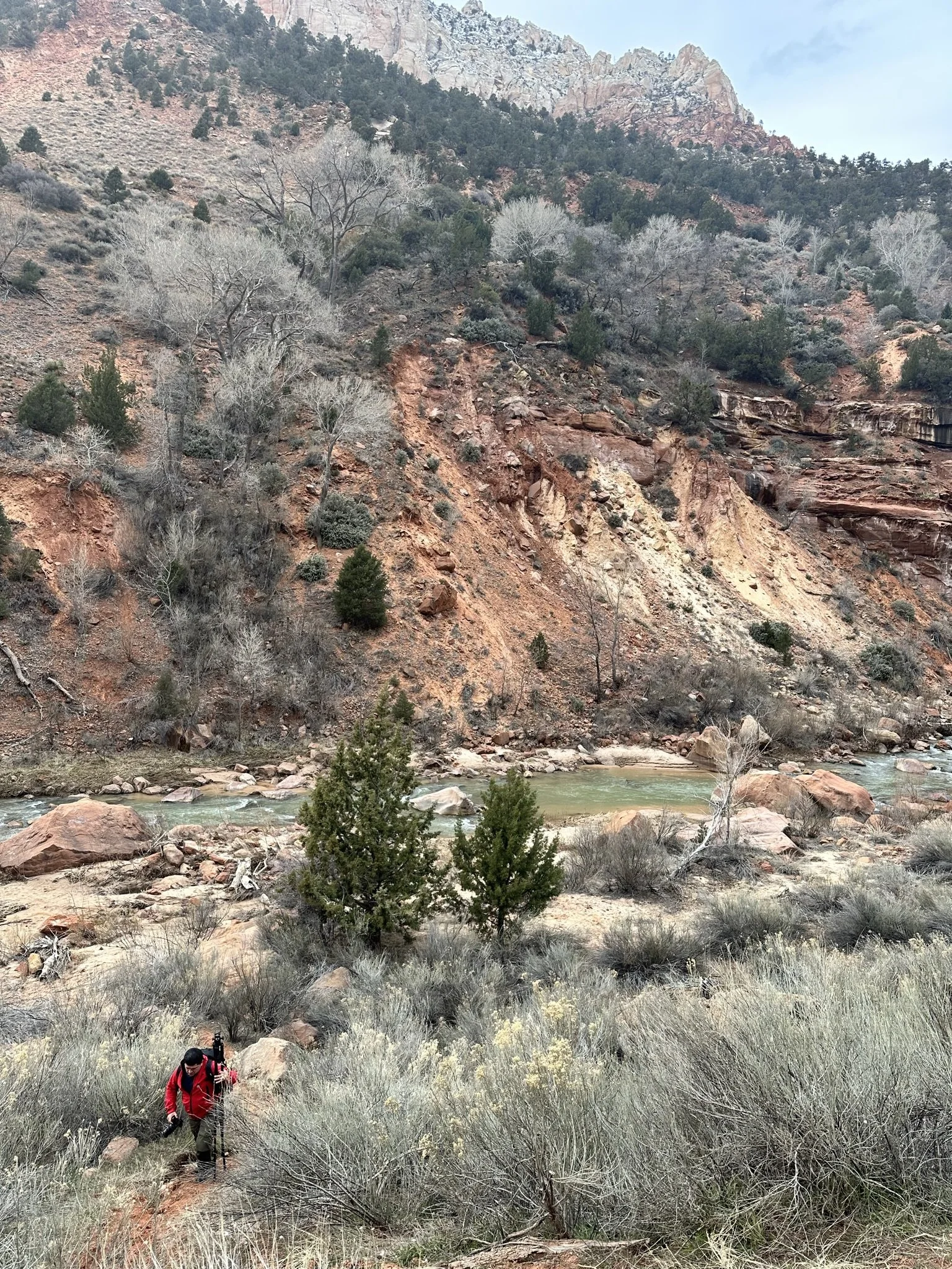 A hiker wearing a red jacket and black pants climbs a trail surrounded by dry bushes and grass, with a rocky river and rugged hillside with sparse trees in the background.