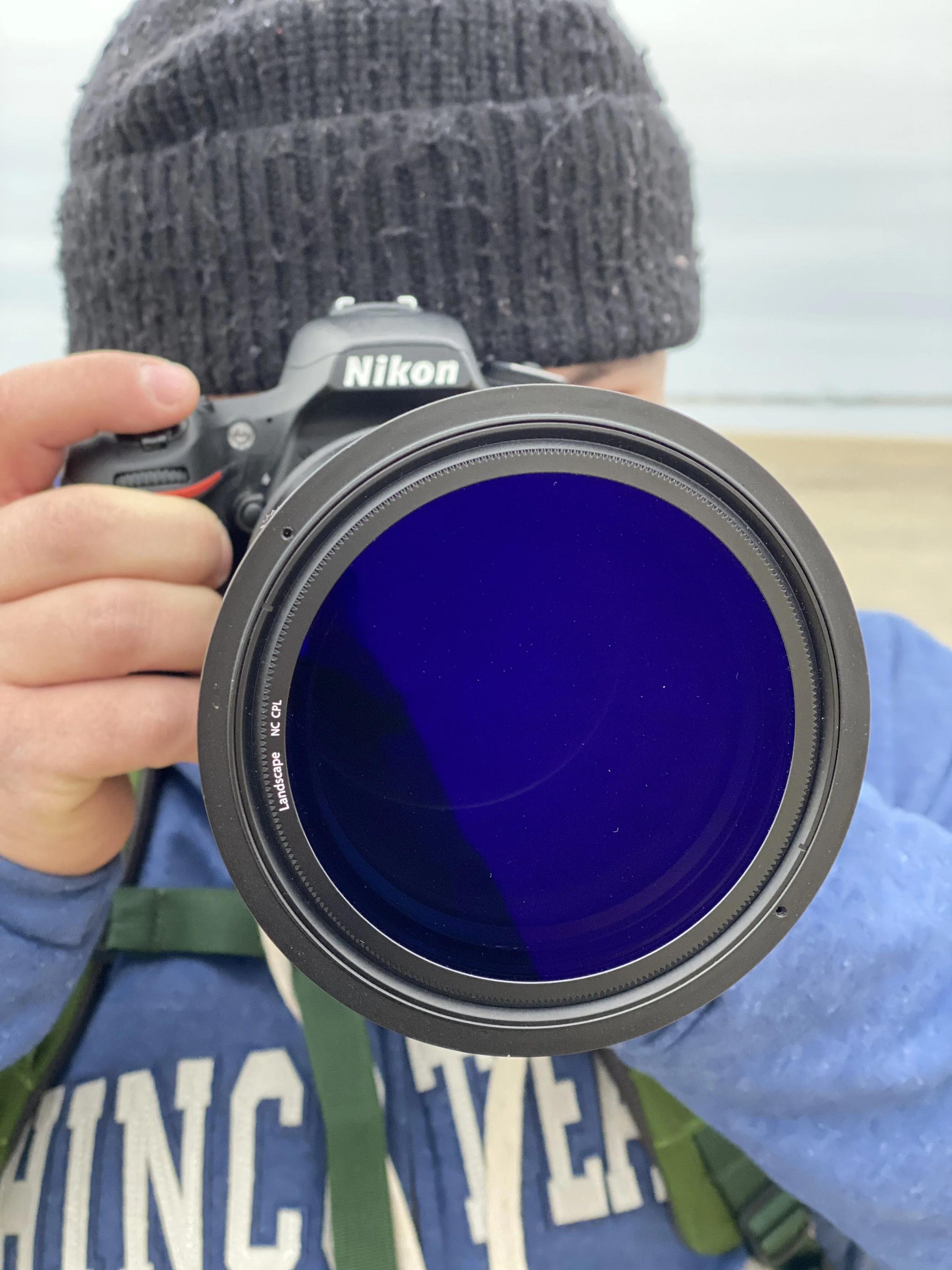 Person holding a Nikon camera with a large telephoto lens, wearing a blue jacket, green backpack, and a black knit beanie, outdoors near a body of water.