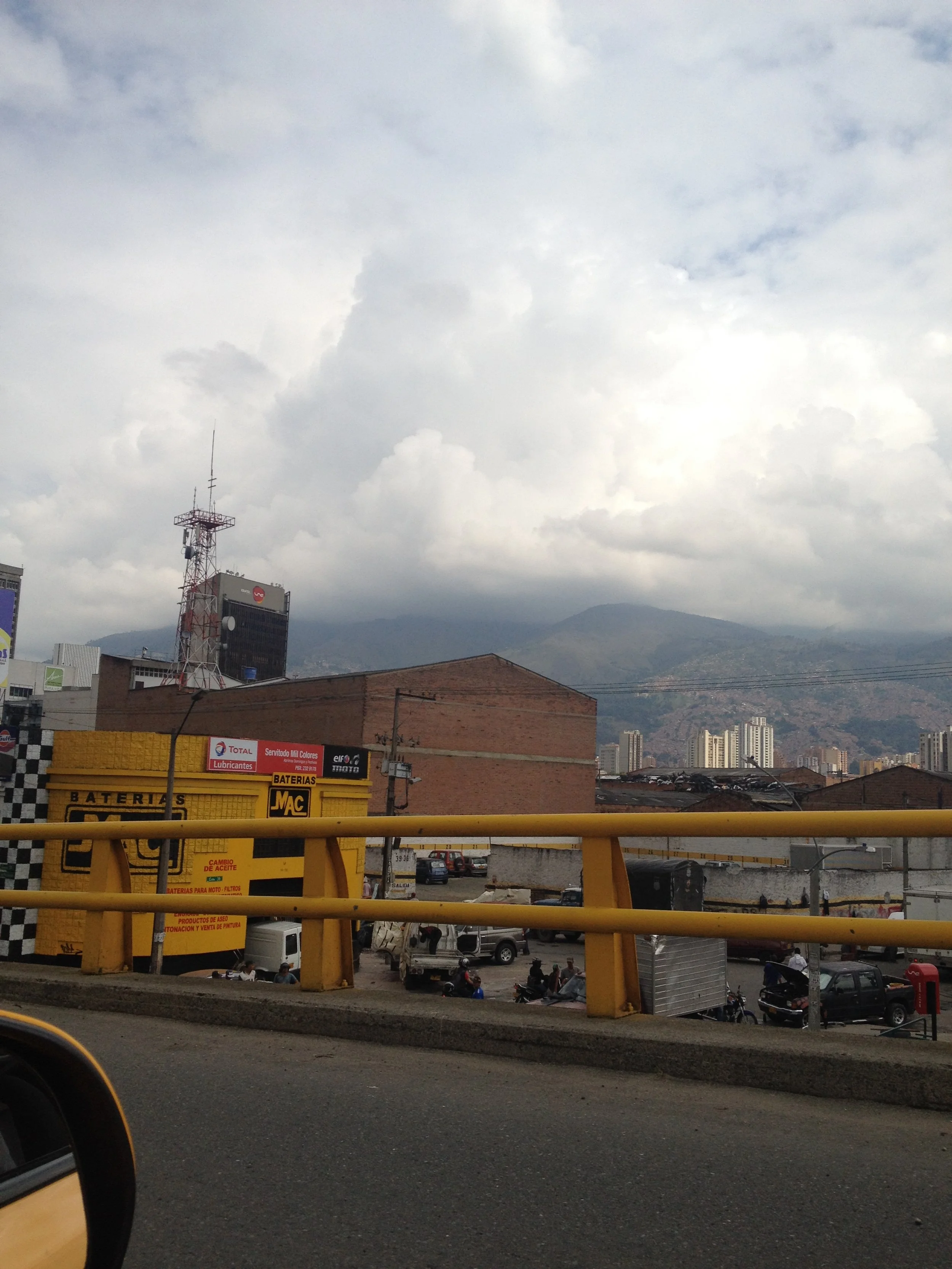 Urban scene with mountains in the background, a cloudy sky, a yellow guardrail, a building with signs, parked vehicles, and city high-rise buildings in the distance.