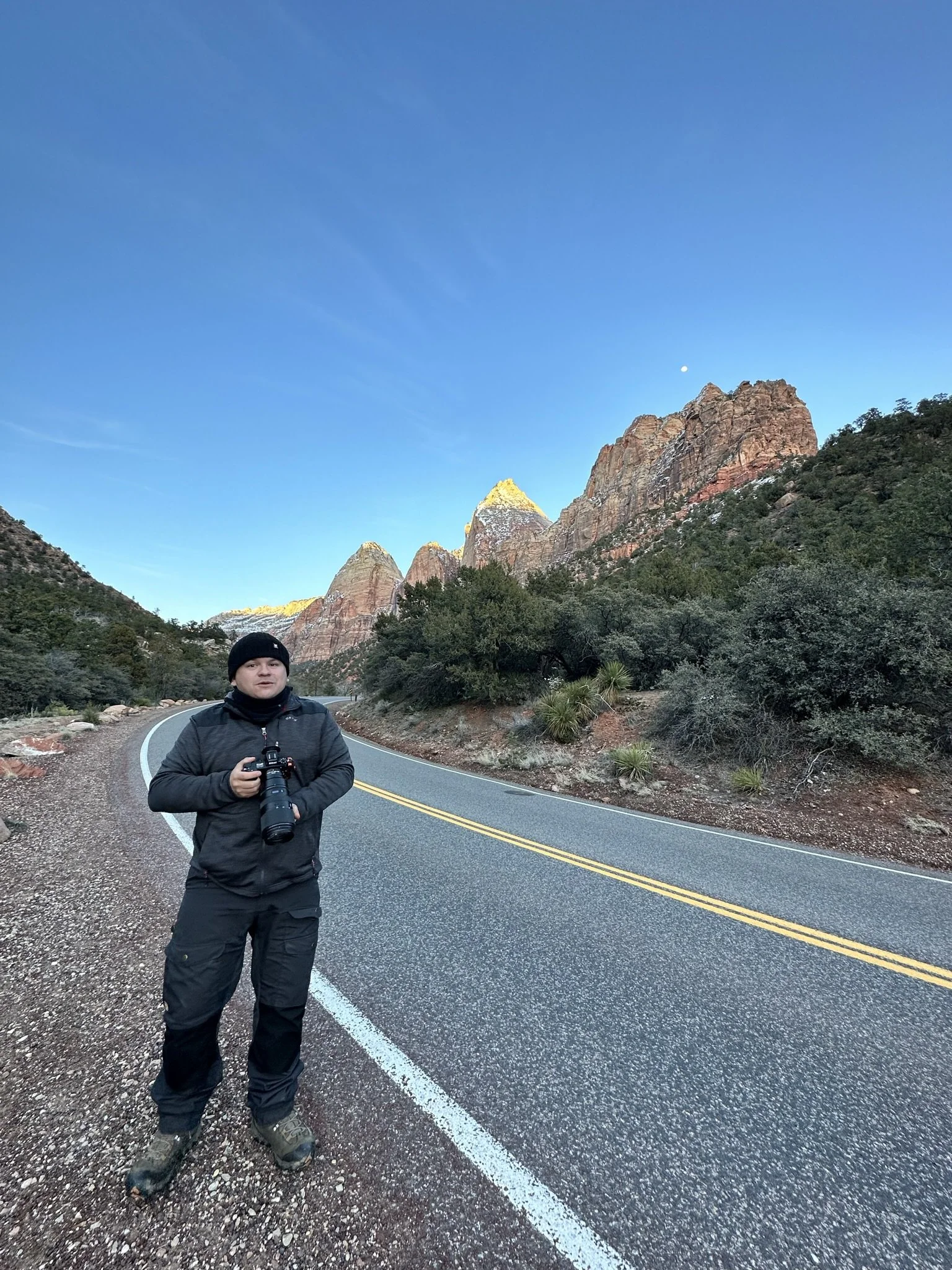 A person dressed in black outdoor gear holding a camera with a long lens, standing on the side of a winding mountain road with red rock cliffs and green shrubbery in the background, under a clear blue sky.