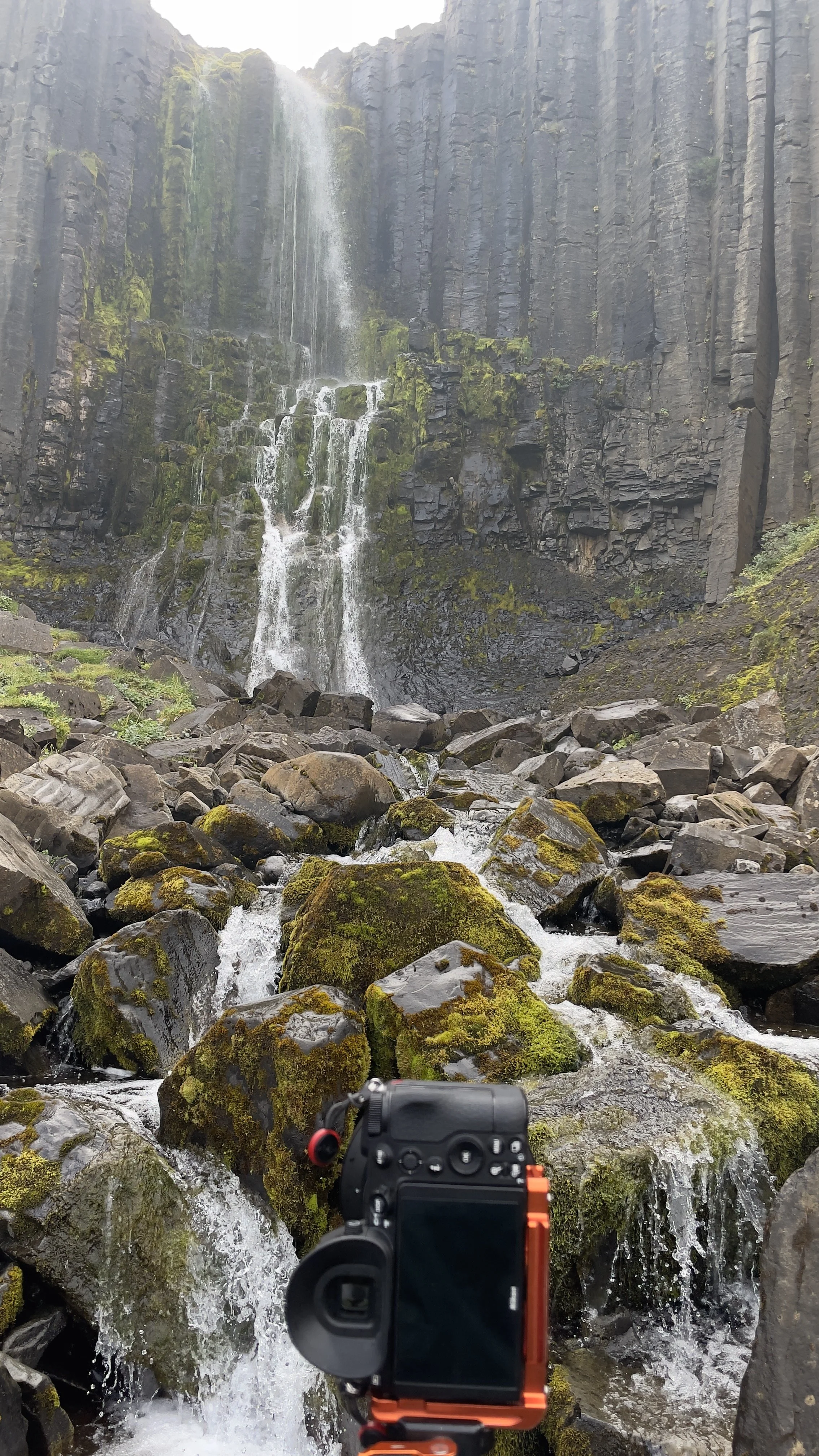 A camera setup on moss-covered rocks in front of a waterfall cascading down a tall cliff in a rugged landscape.